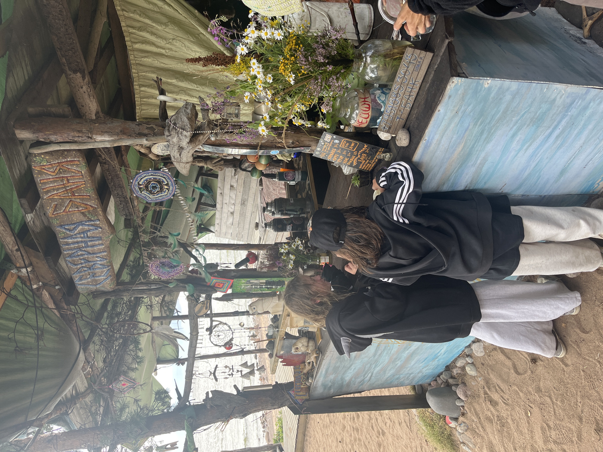The image depicts an outdoor stall with a wooden counter painted in blue and white, situated on a sandy beach. Two individuals, appearing to be teenagers, are standing at the counter, interacting with the items displayed. The stall is adorned with various rustic decorations, including dreamcatchers, wind chimes, and bottles. In the background, a body of water and vegetation are visible, suggesting a natural setting, possibly by a lake or the sea. The overall ambiance suggests a bohemian or artistic market stall.