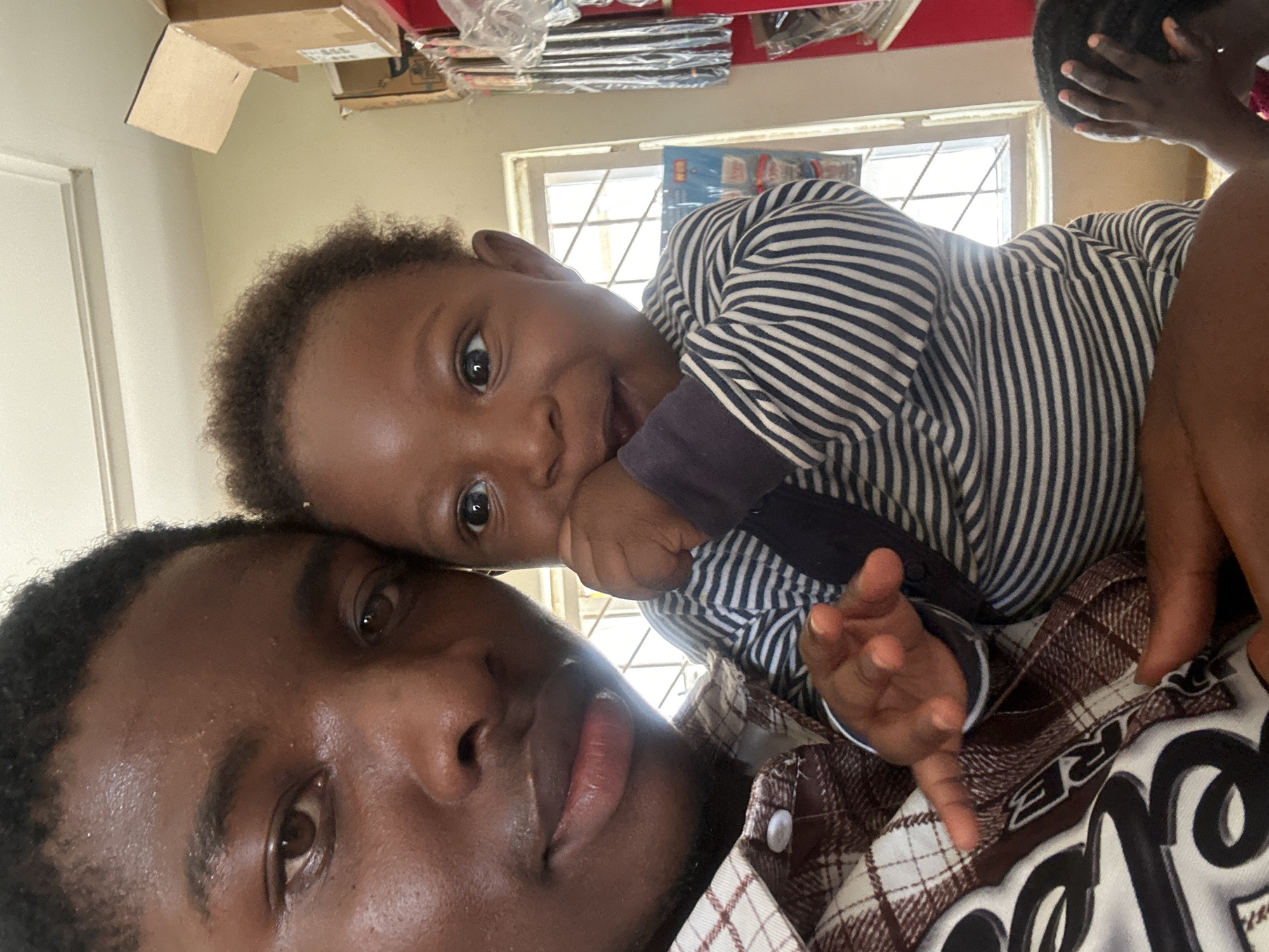The image is a close-up, rotated selfie featuring an adult and a baby indoors, taken in Gura Top, Nigeria.
In the foreground, positioned on the left side of the frame, is an adult male with dark skin, short dark hair, and a faint beard. He looks directly at the camera with a neutral expression, revealing his left eye and full lips. He is wearing a brown and white plaid shirt, with a white button visible on his collar. A partial white-on-black text design, including the letters "RE", is visible on the lower right of his shirt.
Resting above the adult's head, on the upper right side of the frame, is a baby or toddler with dark skin, dark curly hair, and wide dark eyes. The child looks towards the camera with a slightly open mouth and a playful expression, with one hand near their mouth. They are wearing a navy blue and white striped long-sleeved top.
The background shows a window with a grid pattern, through which bright daylight streams, suggesting daytime. Beyond the window, a "LEGO" box or packaging is partially visible. Above and to the left of the window, several cardboard boxes and items wrapped in clear plastic are stacked, indicating an indoor setting that might be a home or a storage area. The intimate framing suggests a shared, warm moment between the two subjects.
