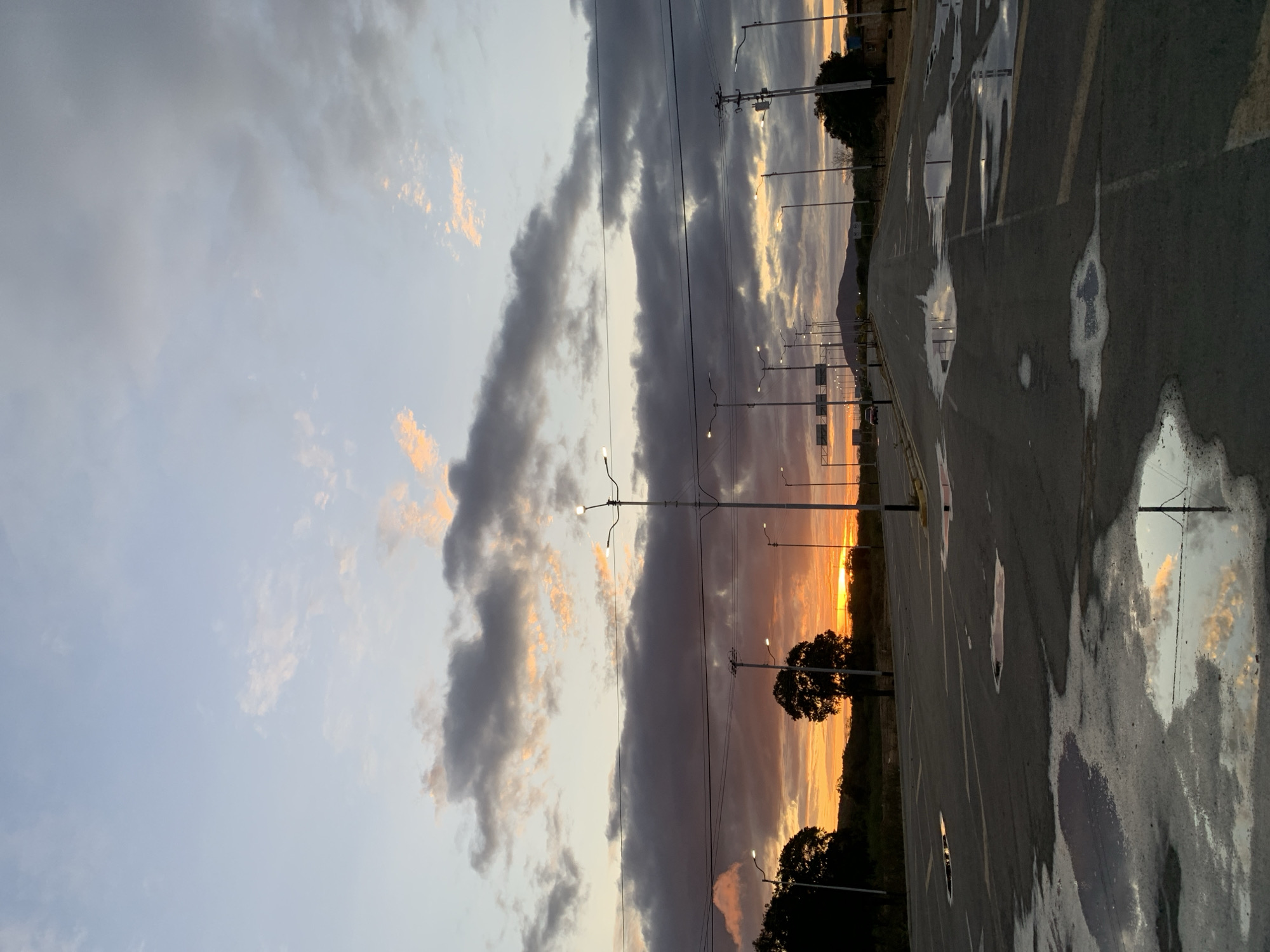 The image captures a serene sunset scene over a wide paved area, possibly a road or large parking lot, in Santa Ana, Venezuela, following a recent rainfall. The sky dominates the upper portion, displaying a dramatic interplay of clouds. Dark, elongated grey clouds stretch horizontally across the middle of the sky, contrasting with patches of vibrant orange and yellow light emanating from the setting sun, which is low on the horizon towards the right. Lighter blue and wispy clouds are visible towards the top left.
The paved surface in the lower half of the image is marked with faint yellow and white lines. Several large puddles are scattered across the asphalt, beautifully reflecting the colorful sky and clouds above. Numerous streetlights, with their bulbs illuminated, line both sides of the road, extending into the distance. Utility poles with overhead wires are also visible against the sky.
On the horizon, dark silhouettes of trees and a subtle, undulating outline of distant hills or terrain are visible, bathed in the warm glow of the setting sun. The time of day is clearly sunset, indicated by the low sun and the rich, warm colors in the sky. The puddles on the ground signify that there has been recent rain. The scene is devoid of people or specific activities, conveying a quiet and contemplative atmosphere. There is no visible text in the image.
