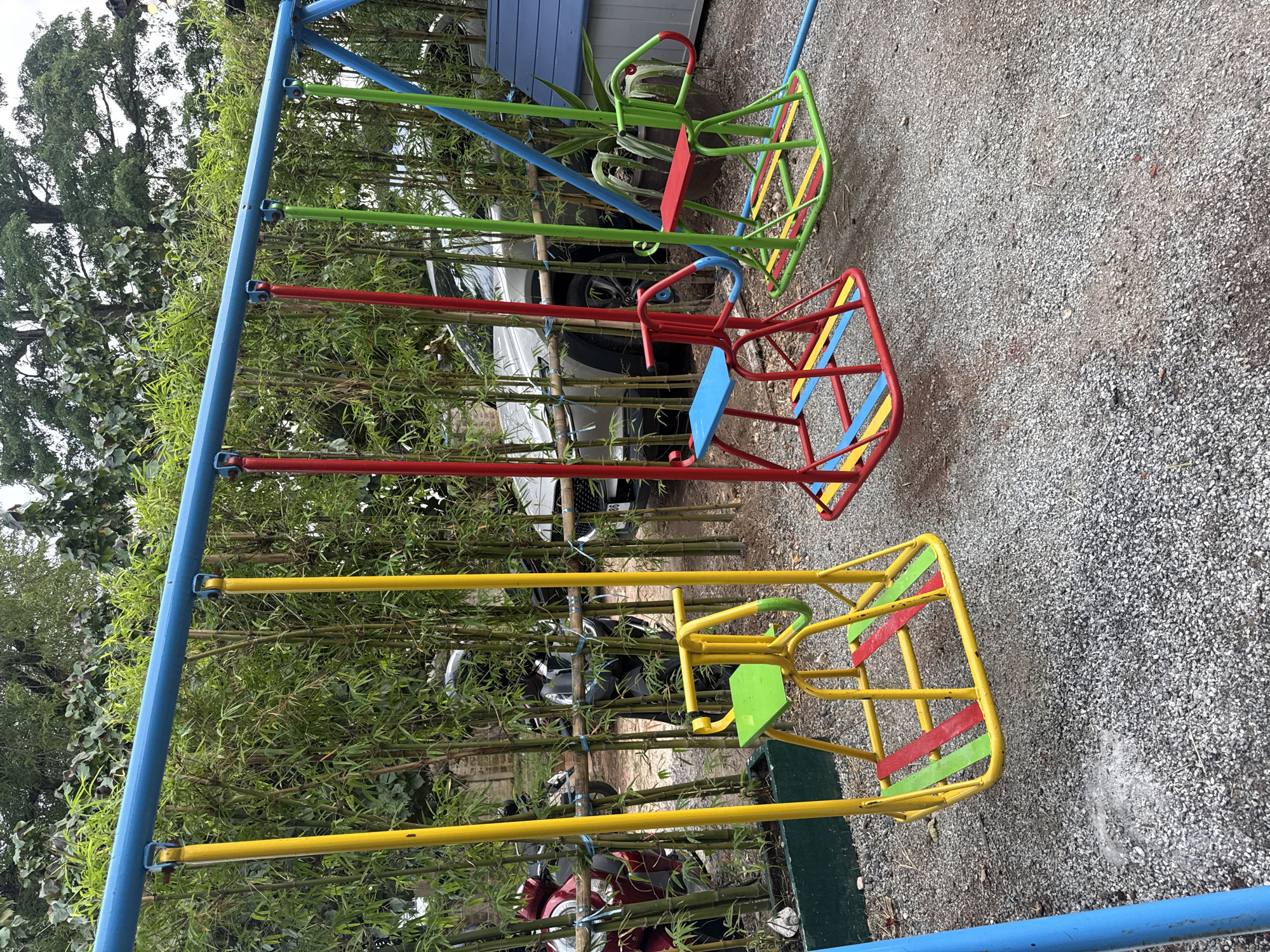 An outdoor playground scene in Wichit, Thailand, features a vibrant blue metal swing set with three colorful swings. From left to right, the swings have yellow support poles, with the leftmost swing featuring a green seat and yellow and red bars on its base. The middle swing has red support poles, a blue seat, and red and yellow bars on its base. The rightmost swing has green support poles, a red seat, and green and yellow bars on its base. The swing set is situated on a ground covered in grey gravel. Behind the swings, a dense bamboo grove is visible, with a light-colored car and two parked motorcycles partially obscured by the bamboo. To the far right of the frame, a small blue and gray playhouse structure is present. The overall environment suggests a casual outdoor recreational area.