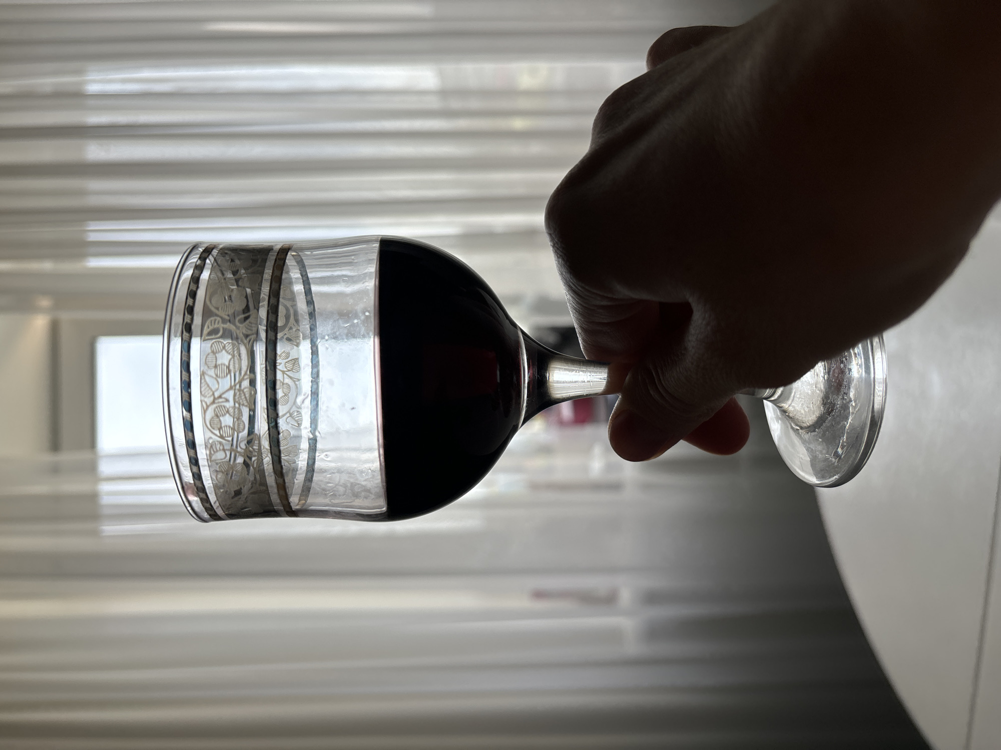 A hand holds a glass of red wine in front of a window. The glass is decorated with floral patterns and silver bands. The background shows white curtains and a window with natural light. The location is unknown.