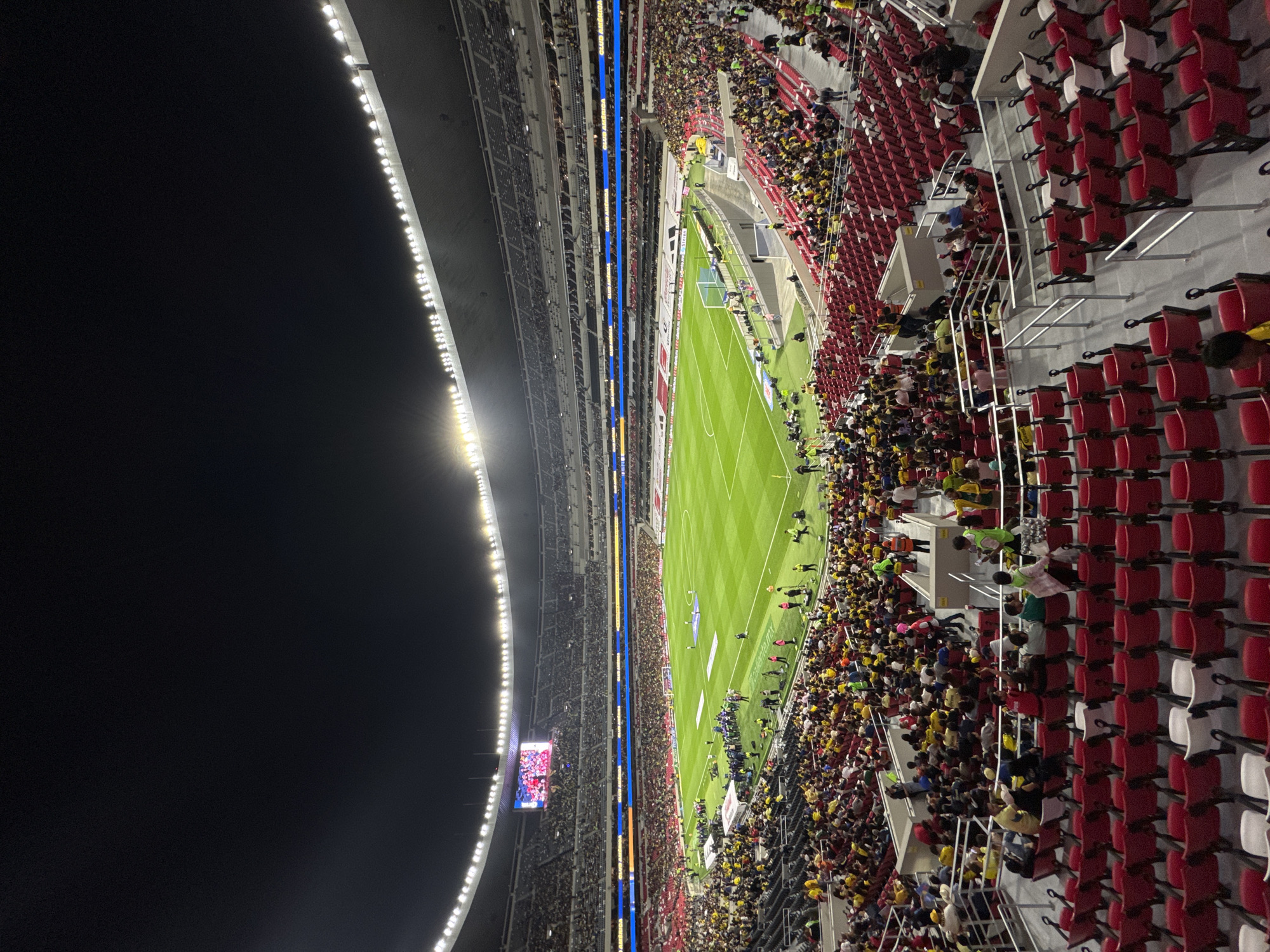 The image shows a soccer stadium in Mexico City, Mexico, filled with spectators at night. The brightly lit green soccer field is in the center, surrounded by stands packed with people, many wearing yellow clothing. Red and white stadium seating is visible on the right side. On the left, there are stadium lights and a large screen displaying an image. There are a large number of people gathered.