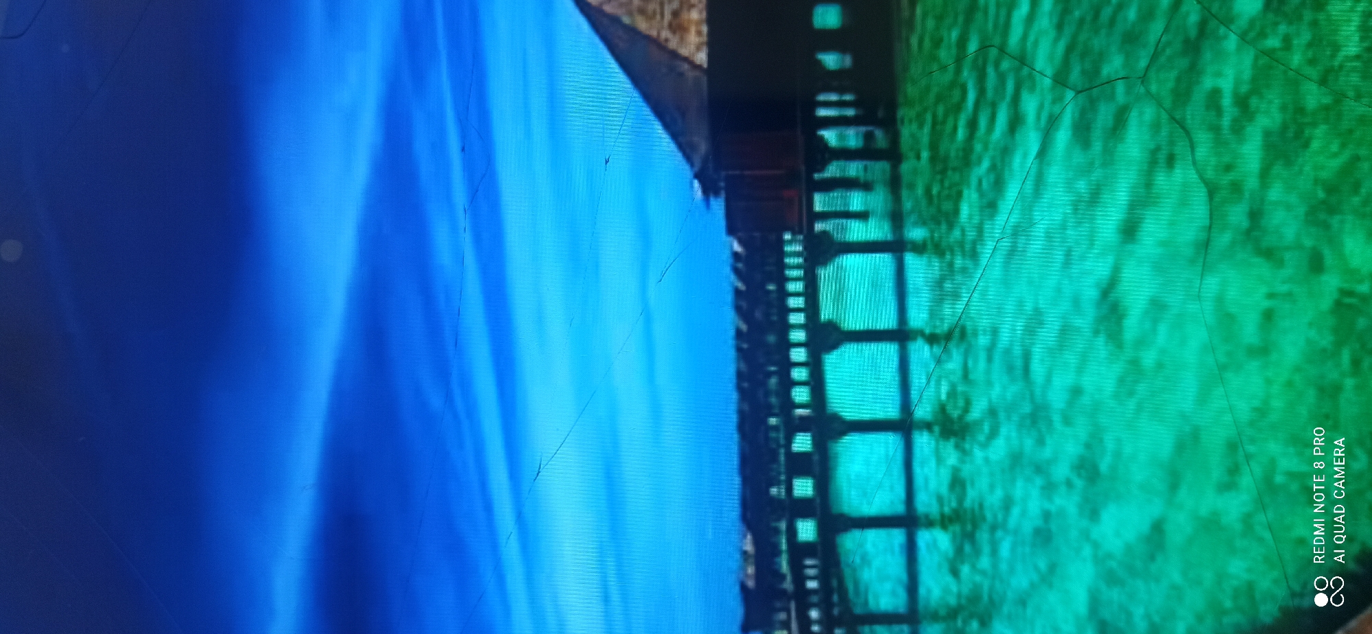 A screenshot shows a vibrant blue sky with wispy clouds above a turquoise body of water. A stilted wooden structure, possibly a pier or walkway, extends across the water, with its reflection visible below. The building appears to have a dark-colored roof and dark wooden walls. The water is clear and shows a rippled texture. The bottom left corner of the image displays text indicating it was taken with a "REDMI NOTE 8 PRO" and "AI QUAD CAMERA." The scene is consistent with a tropical or coastal environment. There is no information about the city of Sedati Agung, Indonesia.