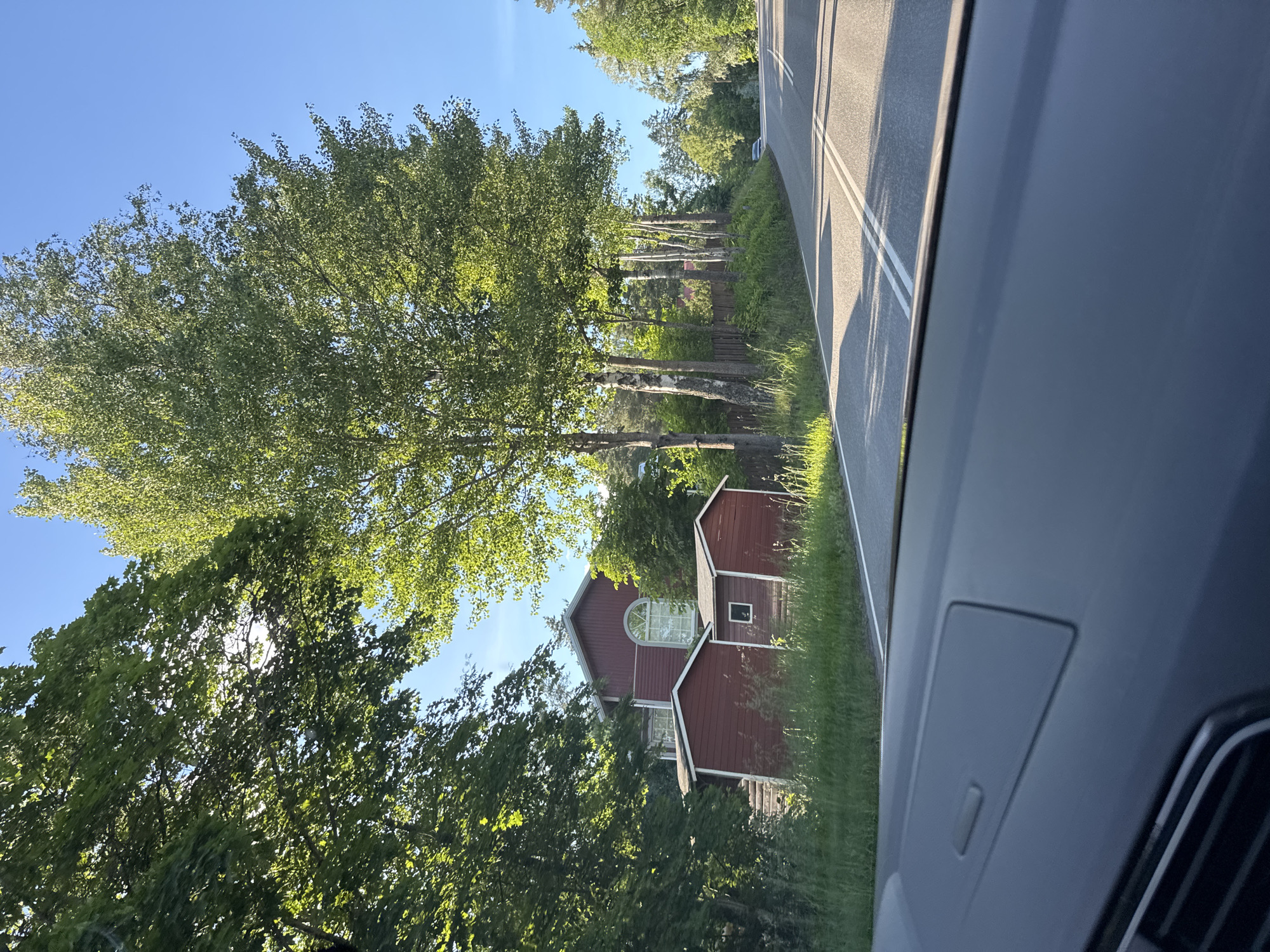 This is a wide shot of a road in a rural area with a house and trees. The image is taken from inside a car. The road has two lanes with double white lines in the middle. The right side of the road is lined with trees, and the left side has a red house with two smaller red sheds in front of it. The house has a large arched window on the second floor. The sky is clear and blue.