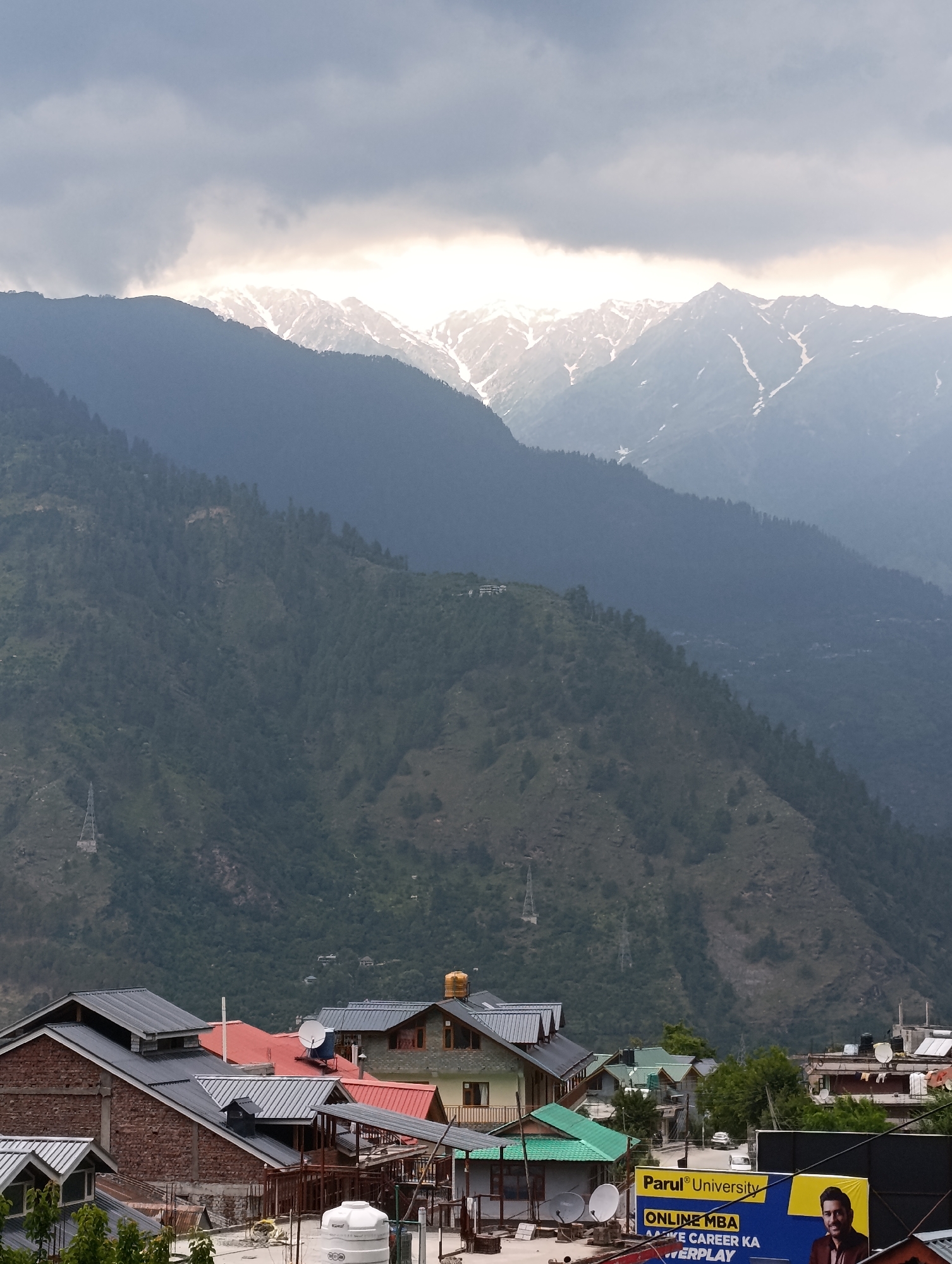 This image captures a layered mountain landscape in Naggar, India, with a town nestled in the foreground under a dramatic sky.
In the background, majestic snow-capped mountains rise, their peaks partially illuminated by sunlight breaking through thick, grey clouds. The light creates a radiant glow along the upper ridges of the distant range, contrasting with the darker, overcast sky above.
Below these towering peaks, a series of densely forested mountains recede into the mid-ground, displaying varying shades of green and blue-grey. Patches of clearings and what appear to be distant structures or buildings are visible on some of the slopes. Faint power transmission towers can also be seen.
The foreground is dominated by the rooftops of several buildings, characteristic of mountain architecture. These houses feature sloping roofs, some dark grey or black, others bright red or green, designed to handle snow. Many roofs have satellite dishes, and white water tanks are visible on some. The buildings are constructed with a mix of brick and plastered walls.
To the bottom right, a large billboard is partially visible. It displays the text "Parul® University," "ONLINE MBA," "AAPKE CAREER KA," and "POWERPLAY," along with the image of a man.
The scene suggests an early morning or late afternoon/evening setting, given the angle of the sunlight and the cloudy conditions. The overall atmosphere is one of serene mountain life under dynamic weather.