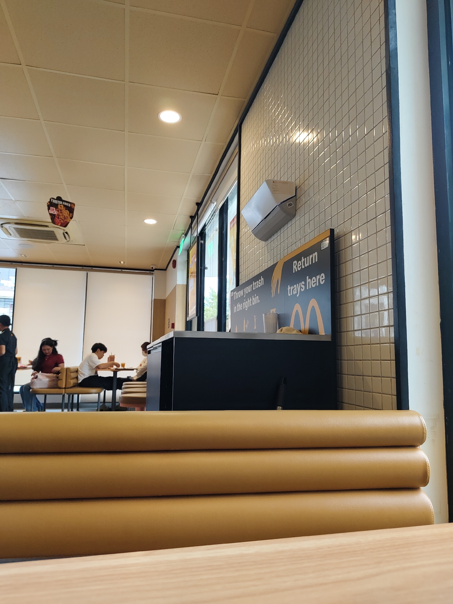 The image shows the interior of a McDonald's restaurant, likely located in Davao City, Philippines, given the context provided. The scene is viewed from a low angle, focusing on a row of padded, mustard-yellow booth seats in the foreground. Behind these seats, a dark counter area is visible with a sign above it that reads "Return trays here" and "Throw your trash in the right bin." The wall behind the counter is covered in small, white, square tiles. Further back, several people are seated at tables, some engaged in eating or conversation. The ceiling is made of acoustic tiles with embedded circular lights. An air conditioning unit is mounted high on the wall in the upper left of the frame. The overall ambiance suggests a casual dining environment.