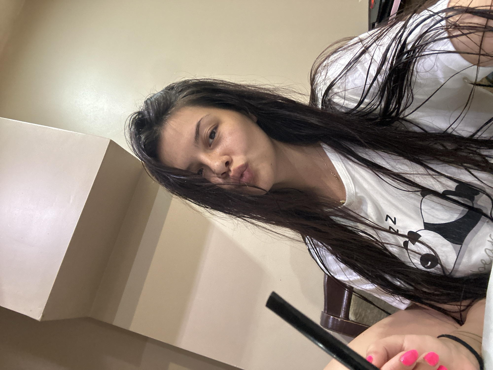 A young woman with long, dark hair and freckles is taking a selfie. She is wearing a white t-shirt with a panda graphic. Her nails are painted neon pink. She is holding a black object, possibly a straw or a pen, up to her mouth and puckering her lips. The background appears to be an indoor setting with a beige wall and a portion of a chair visible. The lighting is warm and diffused. There is no indication of the location being in the city of Sliven, Bulgaria.