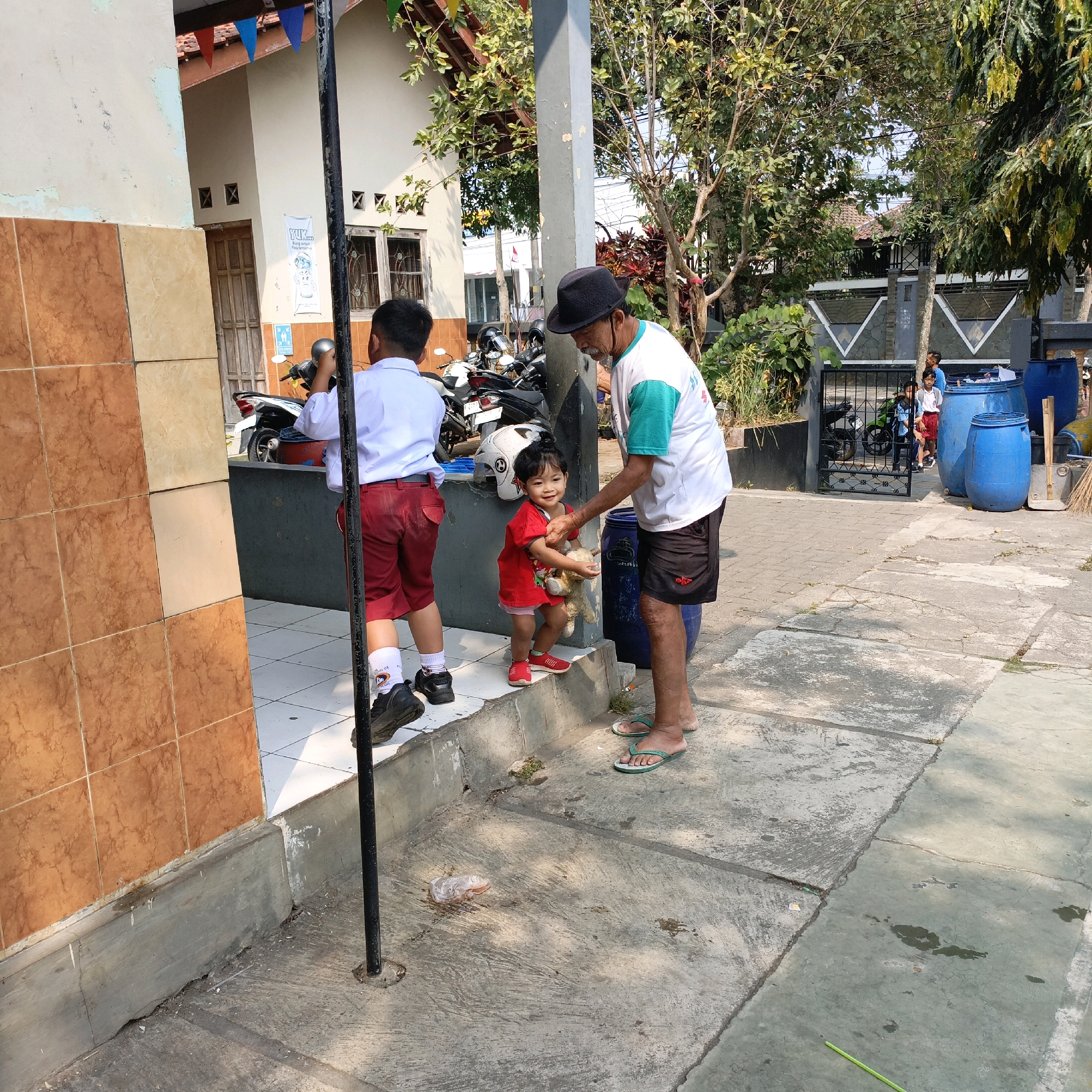 The image depicts a scene outside a building in Salatiga, Indonesia. In the foreground, a boy in a white shirt and red shorts, likely a student, stands facing away from the camera. To his right, an adult man wearing a black hat and a white and green t-shirt interacts with a young child. The child, dressed in red, holds a stuffed animal and appears to be receiving it from the man. Behind them, more children, some in school uniforms, are visible near a black gate. The environment includes a tiled wall on the left, a concrete walkway with some debris, trees, and large blue barrels. The sun is casting shadows, indicating it is daytime.