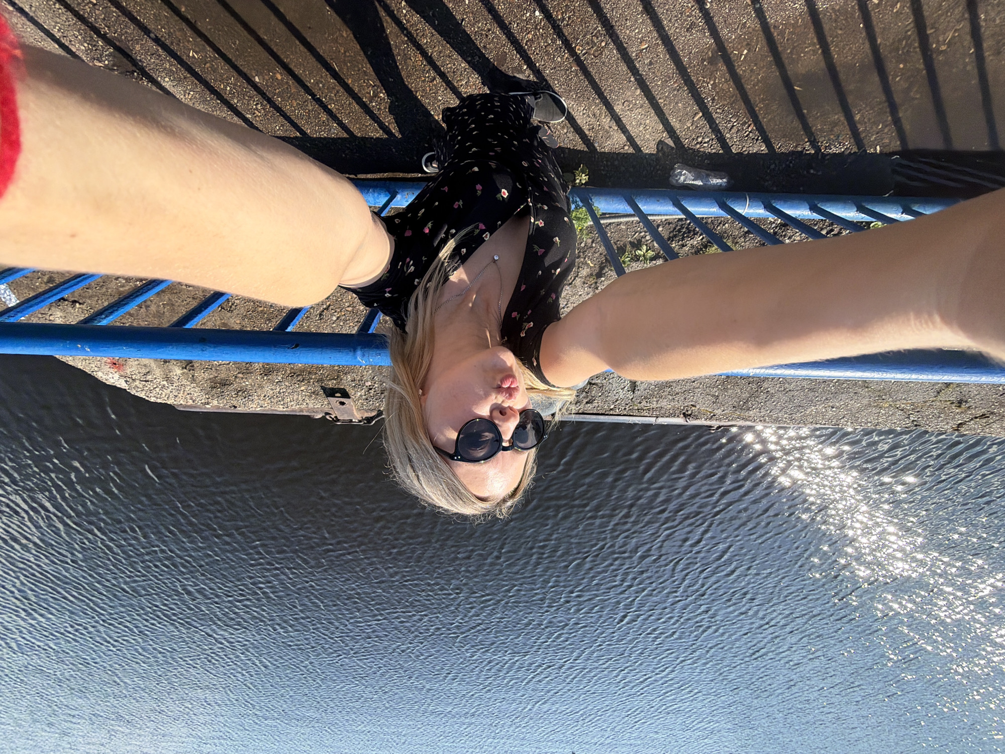A woman with blonde hair and sunglasses is taking a selfie while leaning on a blue railing. She is wearing a black dress with a floral pattern and a necklace. The background is a body of water with sunlight reflecting on the surface. The railing appears to be on a bridge or walkway. The ground is gravelly with shadows cast by the railing. The image is taken from a high angle, looking down. The location is likely the Kama River embankment in Chaikovsky, Russia, given the context provided.