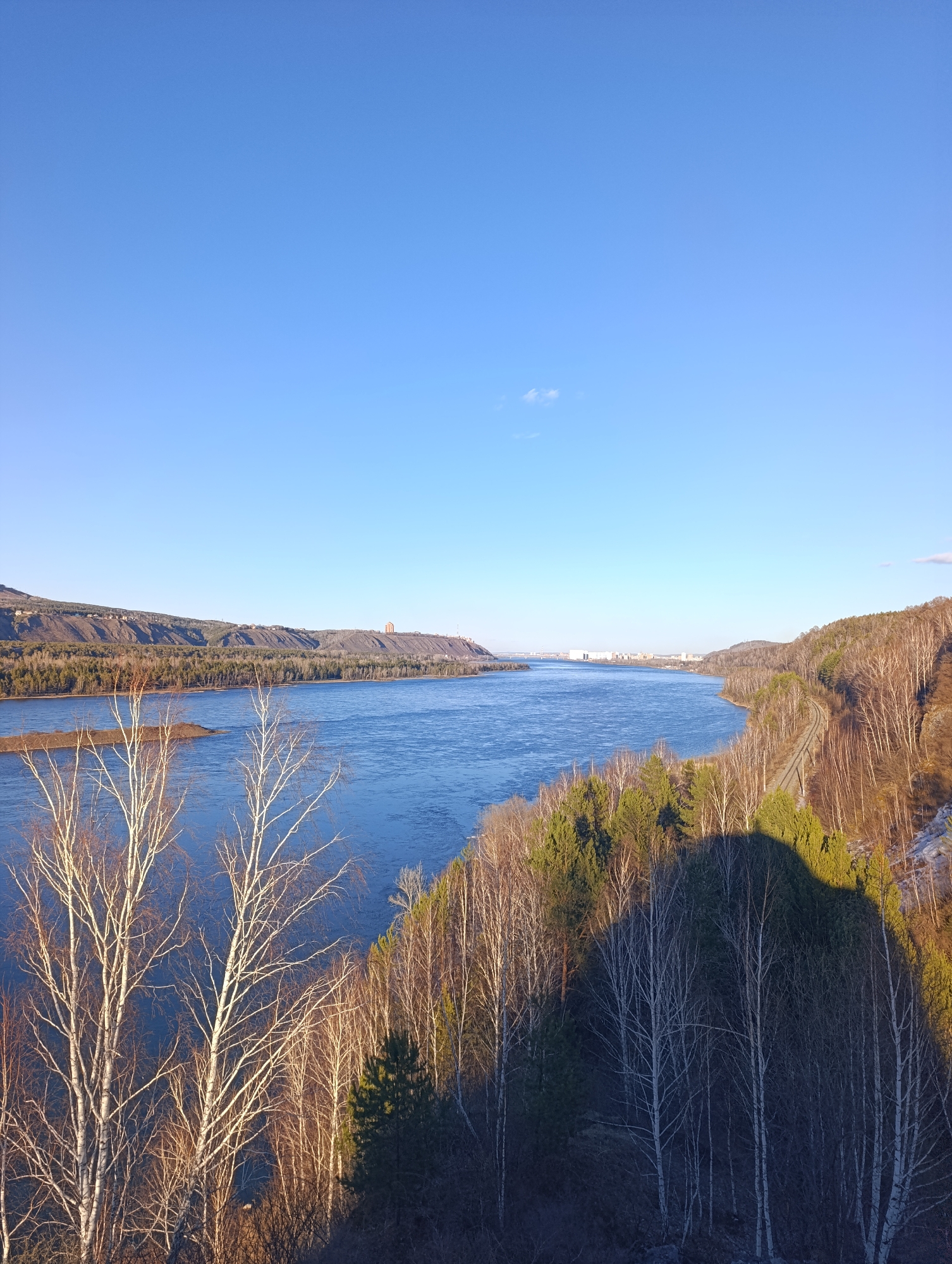 The image shows a wide river flowing through a landscape near the city of Krasnoyarsk, Russia. The sky is clear blue. On both sides of the river, there are forested hills. Deciduous trees with bare branches are prominent in the foreground and midground. Some evergreen trees are also visible. A railway line runs along the hillside on the right side of the river. In the distance, a cityscape can be observed. A distinct shadow is cast on the forest in the lower right corner. A small island is located in the river on the left side.