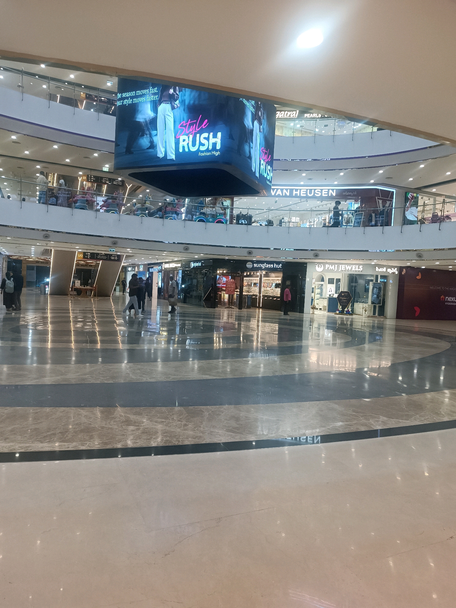 The image depicts the interior of a modern shopping mall in Hyderabad, India. The scene is dominated by a large, multi-level atrium with polished marble floors that reflect the overhead lighting and store signage.
In the center of the frame, suspended from the ceiling, is a large digital display screen showcasing an advertisement for "Style Rush Fashion High" with images of models. Below this, on the first floor, various retail outlets are visible. These include "Van Heusen," "Sunglass Hut," and "PMJ JEWELS," each with distinct signage and storefronts. The stores have glass facades, allowing glimpses of their interiors.
Several individuals are present within the mall. On the lower level, people are walking across the reflective floor. Some are seen on the upper levels, browsing or walking along the balconies. The architecture of the mall is contemporary, with clean lines, multiple levels connected by escalators (partially visible), and a bright, well-lit ambiance. The overall impression is one of a bustling commercial space in a contemporary urban setting.