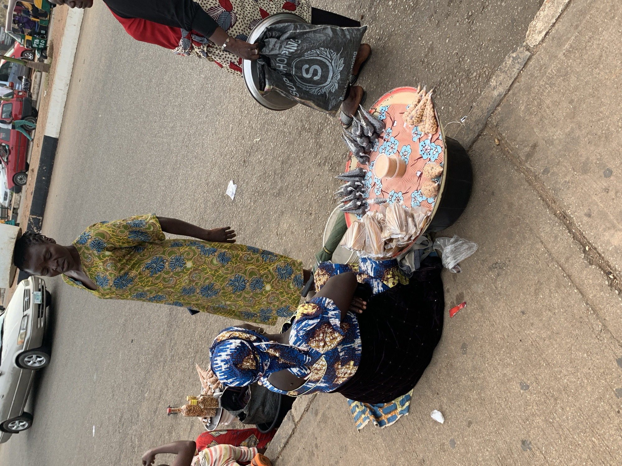 A street scene in Barakin Lerawi, Nigeria, captures a moment of commerce and daily life. In the foreground, a woman sits on the ground wearing a blue and white patterned headwrap and dress. She is facing away from the camera, with her back to the viewer, and appears to be selling goods. To her right, a round, flat surface, possibly a tray or a low table, is covered with a colorful cloth featuring a blue floral pattern. Arranged on this surface are various items, including small, cone-shaped objects that appear to be dried fish, small tubs of what might be spices or condiments, and several small packages of peanuts or similar nuts.
Behind the seated woman, another woman stands wearing a vibrant green and yellow patterned dress. She has a large round object balanced on her head, suggesting she is also transporting goods. Her head is tilted downwards, and her gaze appears to be directed towards the ground.
Further to the right, a third woman is partially visible. She is wearing a black long-sleeved top and a colorful patterned skirt. She holds a black plastic bag with the word "SHOPPING" printed on it in large white letters, along with a circular emblem containing the letter "S". She is also carrying a silver tray.
In the background, a beige sedan is parked on the asphalt road. Other vehicles, including a red car and a motorized tricycle (Keke Napep), are visible further down the street. The environment is an outdoor setting with a paved road and sidewalk, typical of a bustling urban or semi-urban area in Nigeria. The overall impression is one of everyday economic activity taking place on the roadside.