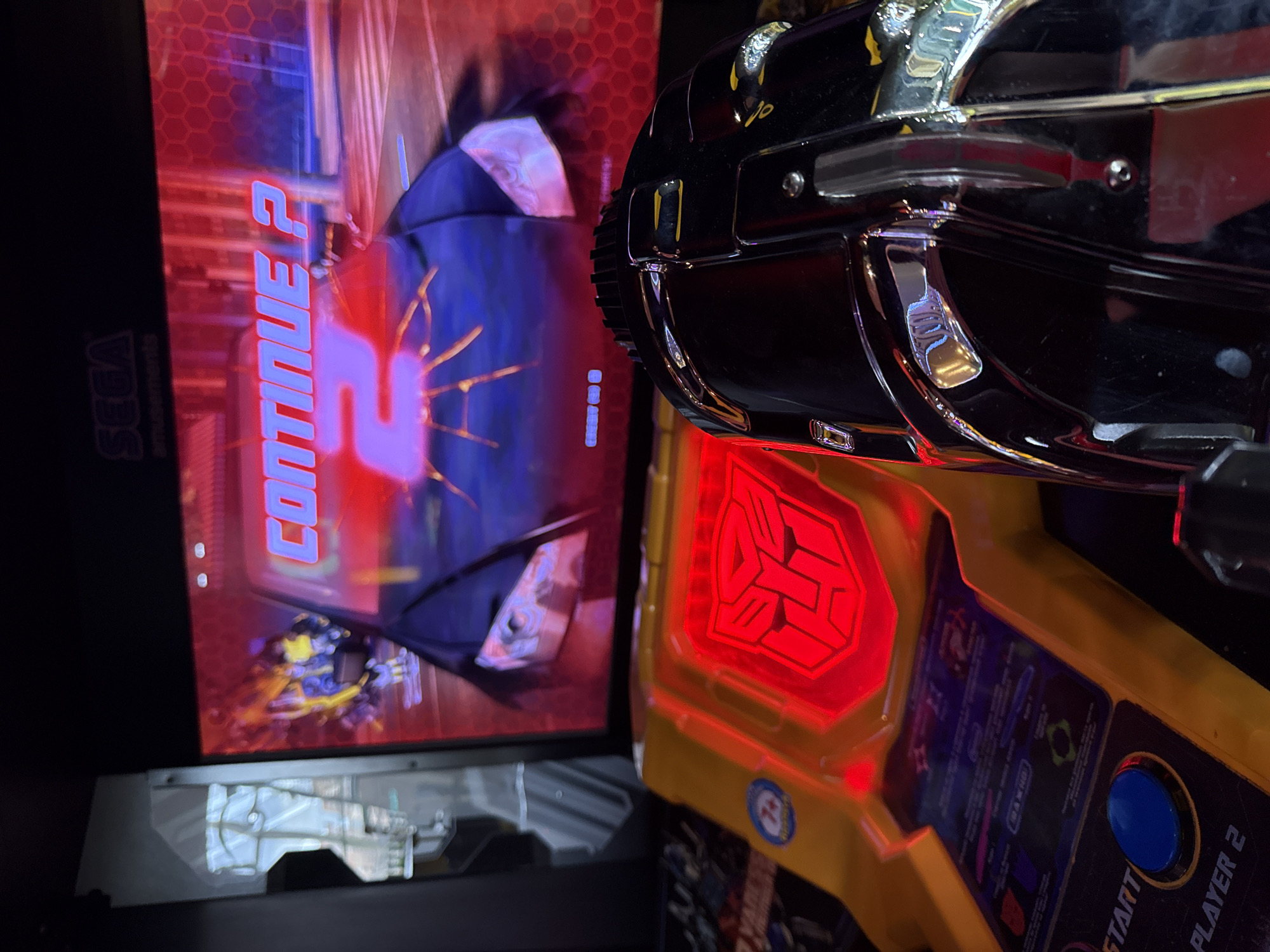 The image shows a close-up of a "Transformers: Human Alliance" arcade game. The screen displays "Continue 2," with a partially visible scene of a Transformers battle. To the right of the screen is the player 2 control panel, featuring a Transformers logo illuminated in red and a yellow panel with the "Start" button visible. The panel also includes a blue button and text indicating "Player 2". Above the panel, there is a chrome-plated gun controller. Based on the game and location, the arcade game is likely located in Glazov, Russia. No people are visible in the image.