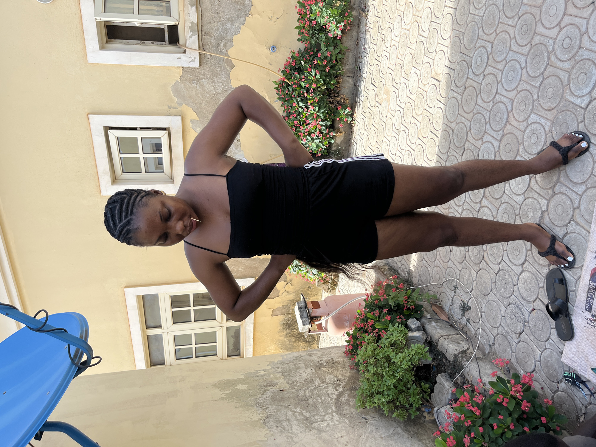 A young woman with braided hair stands on a paved surface. She is wearing a black romper with white stripes on the sides. Her hands are on her hips and she is looking down. The location appears to be an outdoor courtyard area, with plants and flowers growing around the edges. A building with peeling paint and windows is in the background. A gas cylinder sits near a bush with red flowers. There are sandals on the pavement near a white bag. This scene is most likely taken in Abuja, Nigeria.