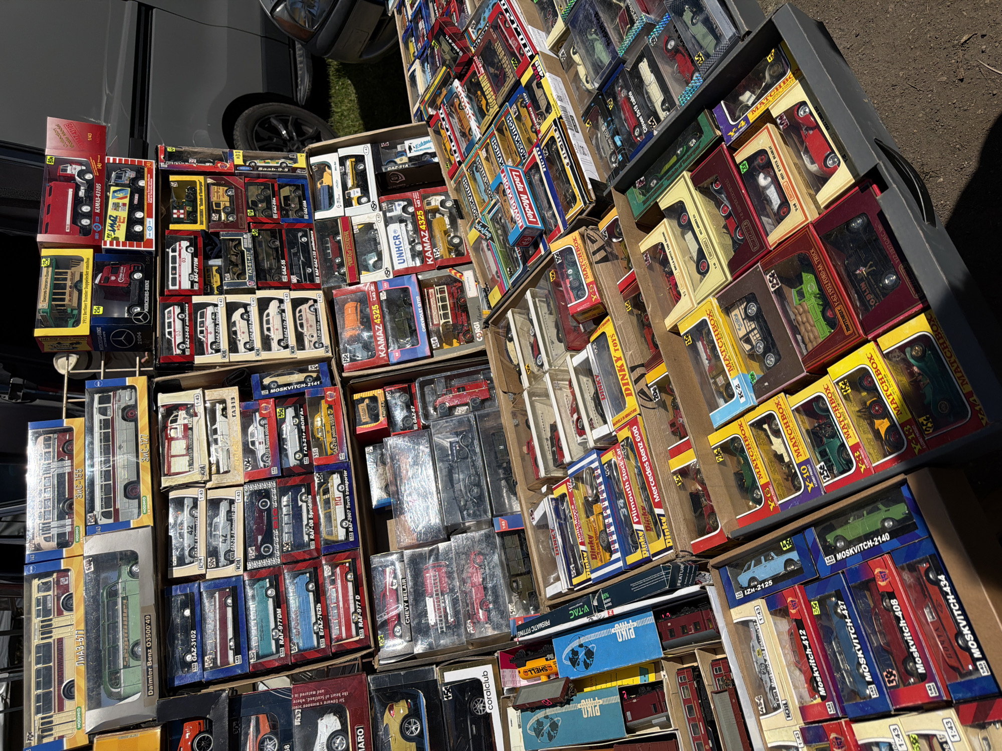 The image captures an outdoor market stall densely packed with numerous die-cast model vehicles, primarily 1:43 scale, displayed in their original packaging or plastic cases. The collection includes a diverse range of Soviet-era and Eastern European transport, such as LIАЗ-677 and ЗиС-155 buses, KAMA3-43105 trucks, and various Moskvitch, GAZ, and RAF passenger cars and commercial vehicles (ambulances, fire trucks). Notable specific models include a green Daimler Benz O3500'49 bus, a 1911 Berliner Daimler Doppeldecker Omnibus, and a Chevrolet Belair. Brands like Matchbox, Dinky, Modimio, and AVD Models are visible. The models are arranged in stacked cardboard and wooden crates on tables, indicating a retail setup. The ground beneath the stall is dirt or gravel. In the background, a dark-colored commercial van with a visible wheel and a portion of a silver passenger car are parked under bright natural sunlight. The scene is consistent with the general appearance and merchandise found at the Latgale Market (Latgalīte), a prominent flea market located in the city of Riga, Latvia. No individuals are visible within the frame.