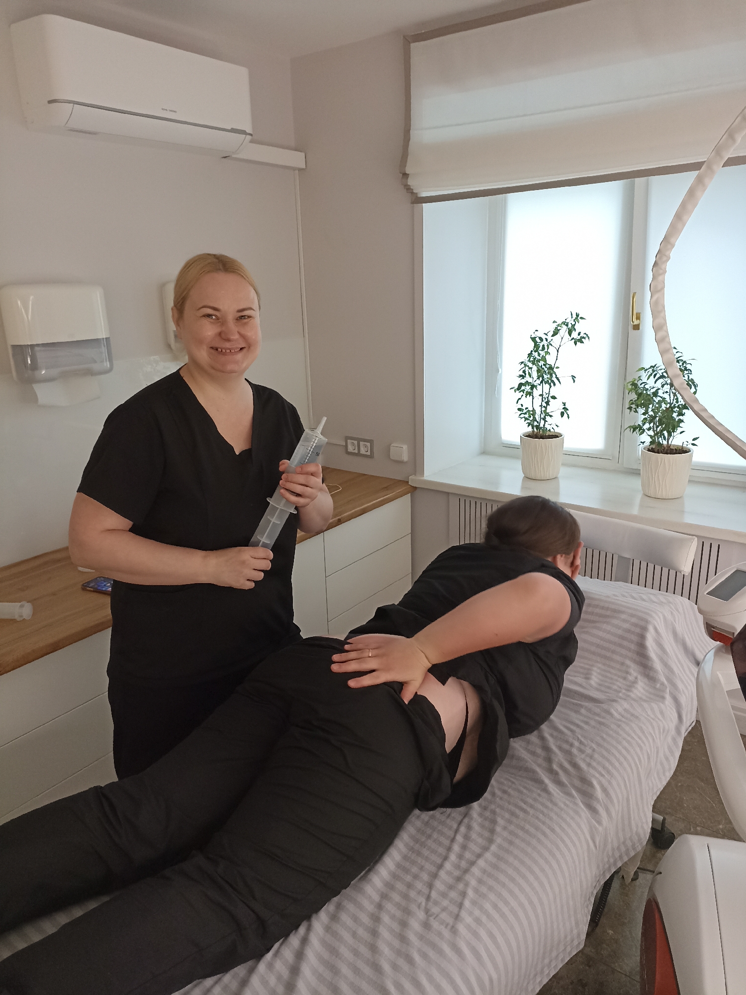 Here is a detailed description of the image:
**Scene:** The image depicts an indoor scene, likely a medical or beauty clinic, in Votkinsk, Russia.
**People:** Two women are visible. One is standing, facing the camera, and wearing a black uniform. She is holding a large, clear plastic syringe. She has blonde hair and is smiling. The other woman is lying face down on a treatment table. She is also wearing black clothing and has her hand placed on her lower back.
**Objects:** The scene contains various objects including: a treatment table with a striped cover, a large plastic syringe, an air conditioning unit on the wall, a paper towel dispenser, a wooden countertop, an electrical outlet, a window with two potted plants, a beauty device, and a small table.
**Actions and Interactions:** The standing woman is holding the syringe, possibly preparing for a treatment. The woman on the table appears to be receiving or preparing for a medical or cosmetic procedure. She has her hand on her lower back.
**Environment:** The environment is clean and well-lit, suggesting a professional medical or beauty clinic setting. The presence of potted plants adds a touch of nature to the sterile environment.