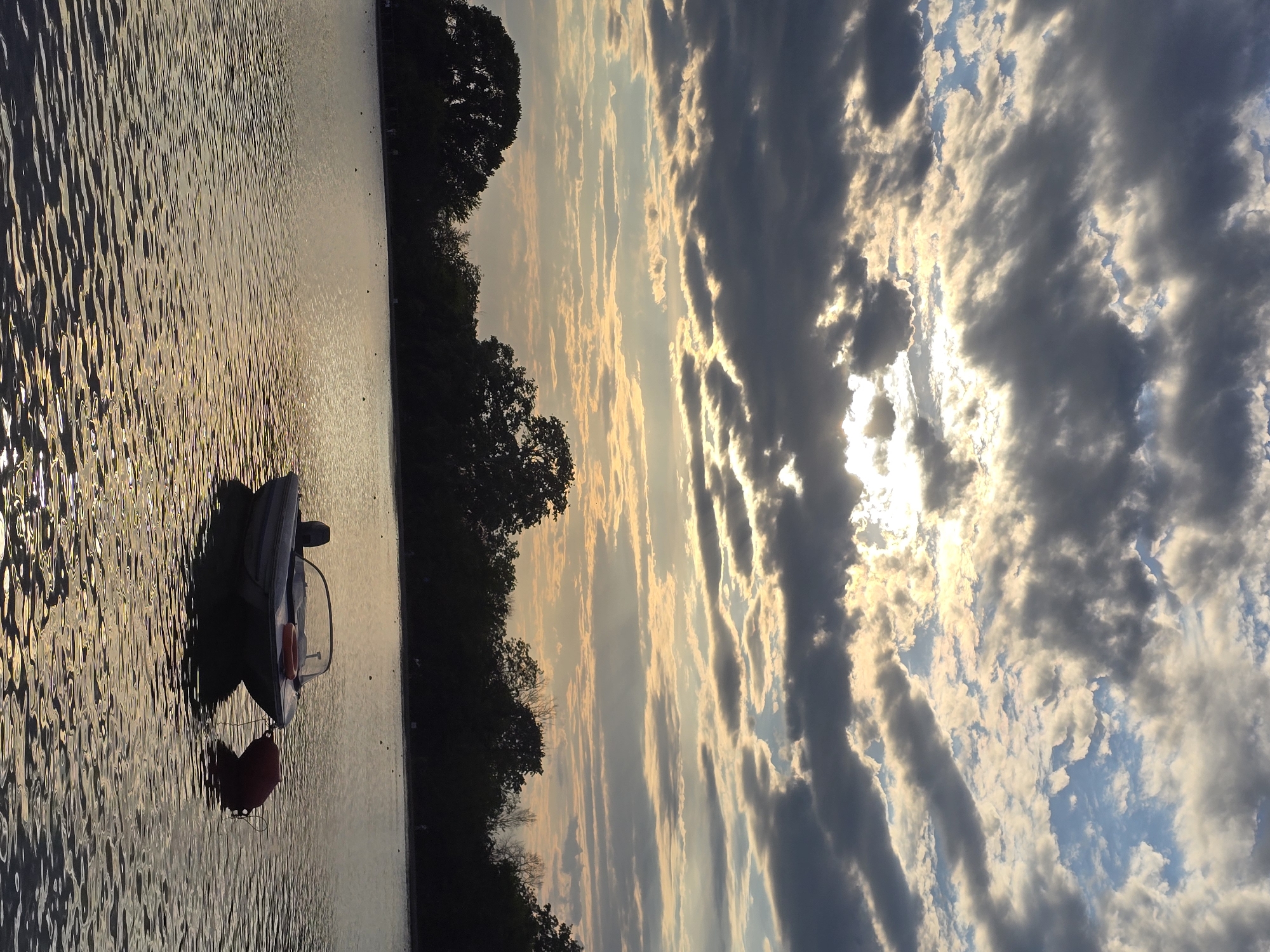 The image shows a small boat on a body of water near a treeline. The sky is filled with clouds, and sunlight is visible breaking through them. There is a red buoy near the boat. The city and state are unknown.