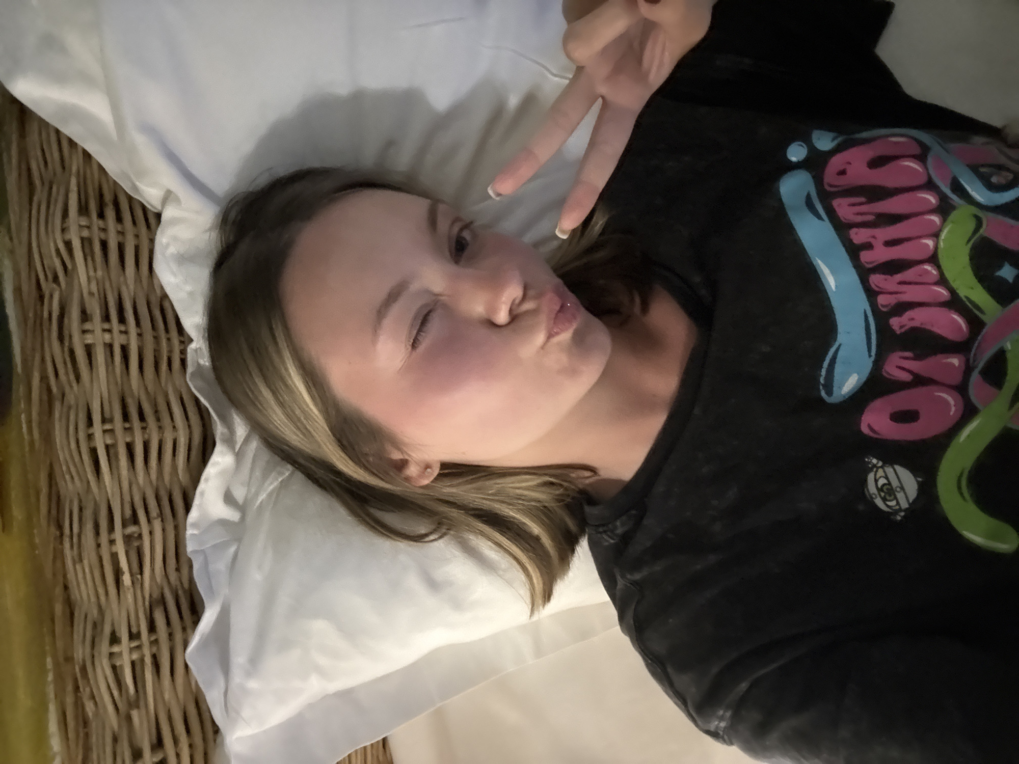 A woman is lying on a bed with white sheets and a pillow, making a peace sign with her right hand and winking her left eye while pursing her lips. She wears a dark-colored T-shirt with a graphic design. A wicker headboard is visible behind the pillow. Based on the prompt the location is somewhere in Nha Trang Ward, Vietnam.