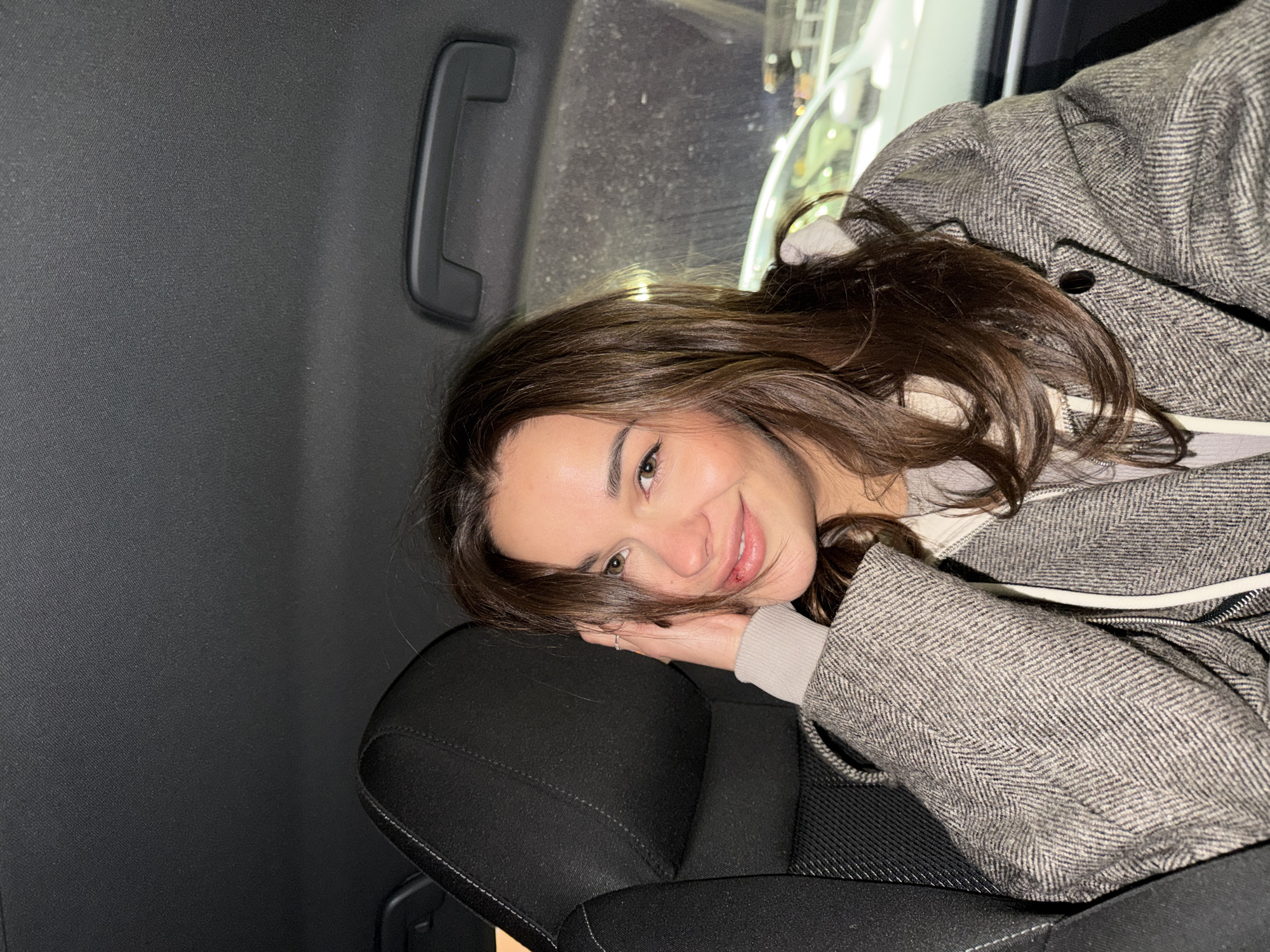 The image, rotated counter-clockwise, features a young woman smiling directly at the camera from inside a vehicle, likely in Krasnoyarsk, Russia. She has long, wavy dark brown hair and light-colored eyes. Her head is gently resting on her right hand, which in turn is propped on a dark, textured fabric car seat. She wears a grey tweed or herringbone patterned coat over a lighter, possibly hooded, inner top with a visible white zipper.
The background reveals the interior of the car, with dark grey paneling and a black overhead grab handle visible in the upper left. Through a window in the upper right, blurred lights and shapes suggest it is nighttime outside. The overall lighting is dim, emphasizing her friendly and relaxed expression. No visible text is present in the image.