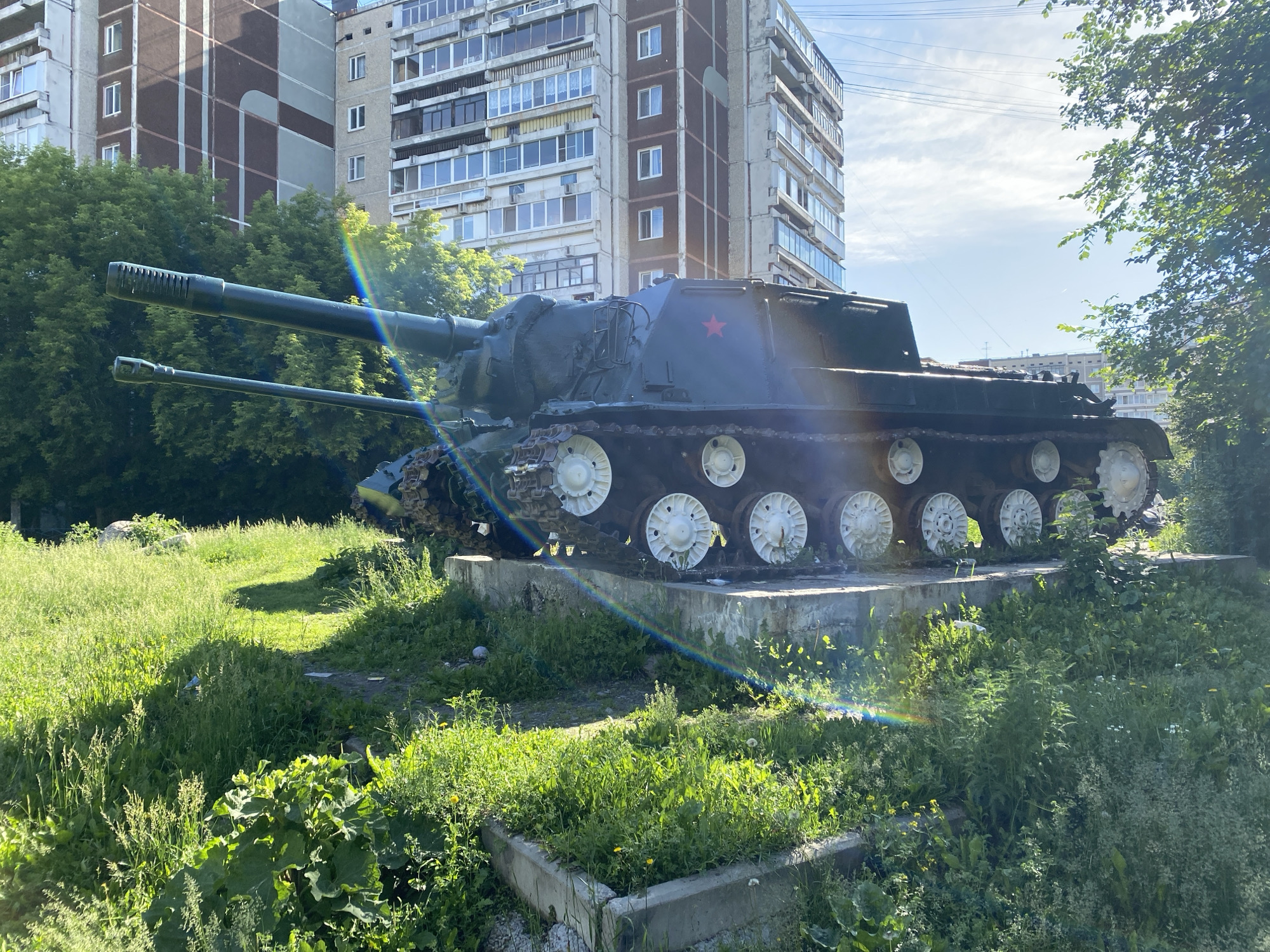 This image depicts a Soviet-era tank, possibly a self-propelled gun, displayed as a monument. The tank is painted dark green and features a prominent red star on its turret, indicating its Soviet origin. It is mounted on a concrete pedestal surrounded by lush green grass, weeds, and some small trees. In the background, two multi-story apartment buildings are visible, suggesting an urban setting. The sky is blue with scattered clouds, and sunlight casts shadows, creating a bright, outdoor scene. A faint rainbow effect is visible due to lens flare. The tank appears to be a static exhibit, likely in a public space or park. The specific location is identified as Yekaterinburg, Russia, near one of the city's housing complexes. There are no people or animals visible in the image.