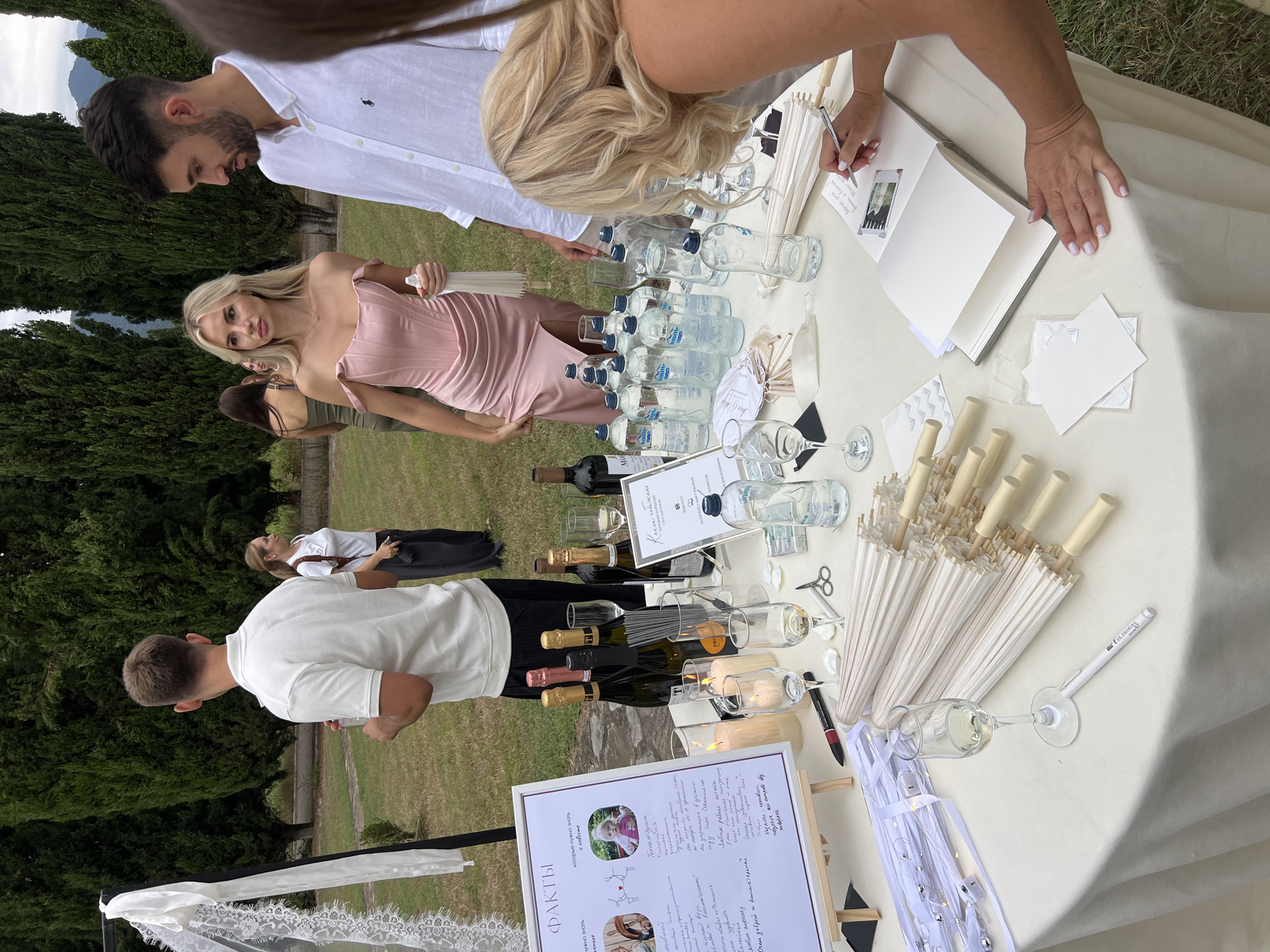 In Saniore, Georgia, an outdoor event is taking place with a round table set up for guests. The table is covered with a cream-colored tablecloth and displays various items. There are several bottles of water, some wine bottles, champagne bottles, and champagne flutes. Small white umbrellas are neatly stacked on one side of the table, and a guest book is open, with a woman with blonde hair writing in it with a pen. Candles in glass holders are also present, along with a sign with text and illustrations, and a pair of scissors. In the background, several people are milling around in a grassy area with tall evergreen trees lining the perimeter.