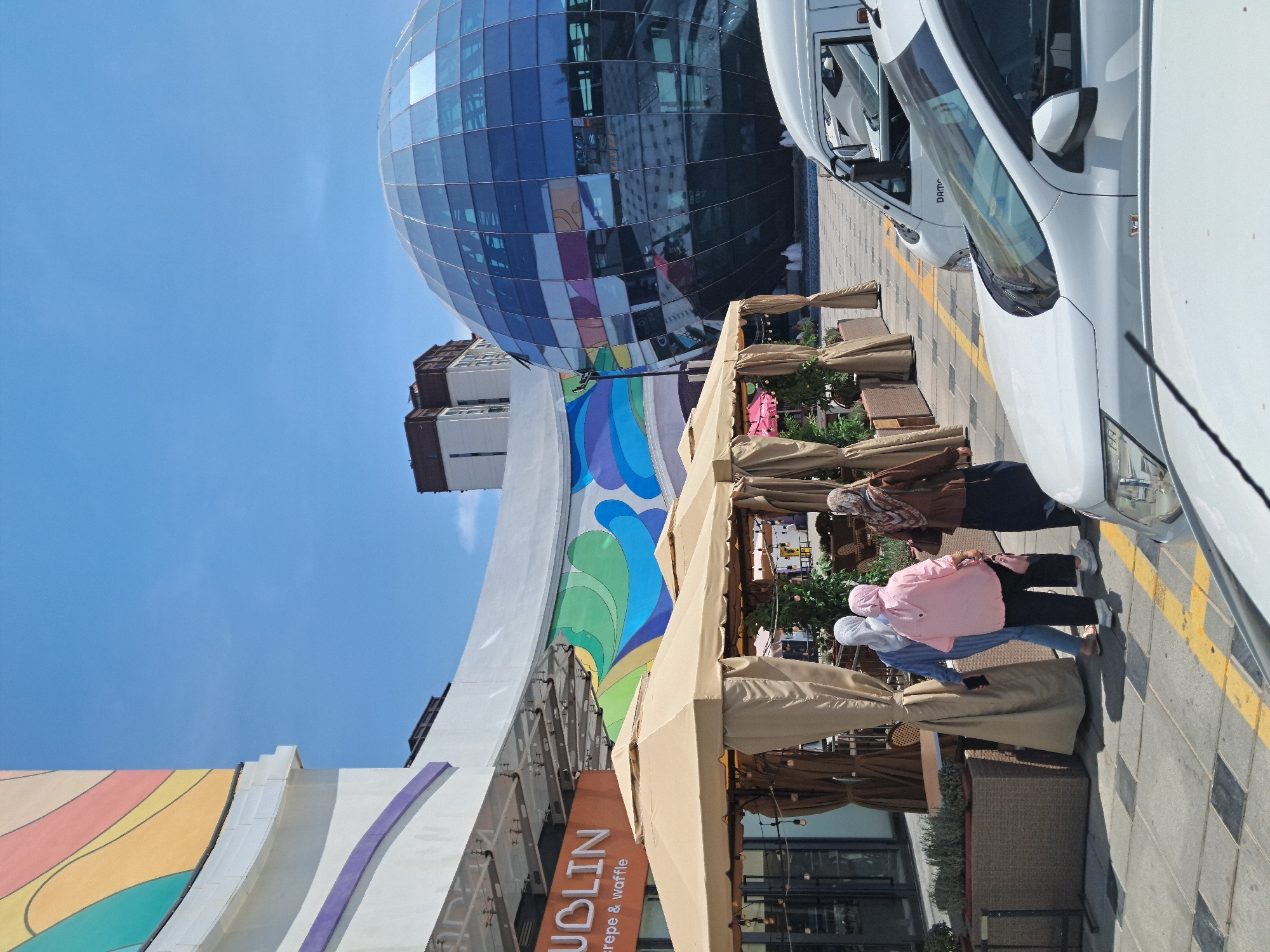 In a city setting under a clear blue sky, a row of beige outdoor dining tents stands on a paved walkway. Three women wearing hijabs are walking away from the camera, two in front and one behind, in the vicinity of the tents. A large, spherical building with a glass facade, reflecting the surrounding environment, is visible in the background, along with other modern buildings. Several white vehicles are parked on the walkway. A sign with the word "DUBLIN" in orange is displayed above the tents.