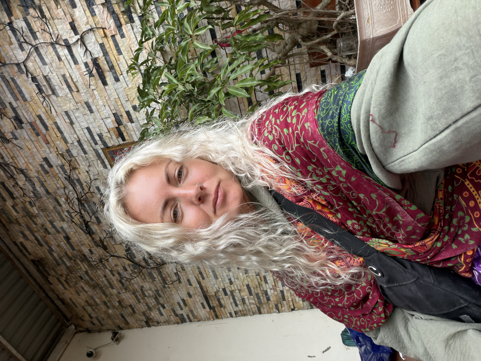 A young Caucasian female with long, wavy blonde hair is seated outdoors. She is wearing a light green hoodie with a decorative red and green patterned scarf draped over her shoulders. A black crossbody bag is positioned in front of her. She has a nose ring and is looking directly at the camera. The background features a wall constructed of textured, multi-colored rectangular stones, with dark, branch-like decorative elements on its surface. To the right, a lush green plant in a pot is visible. The scene appears to be in Hướng Phùng Commune, Vietnam.