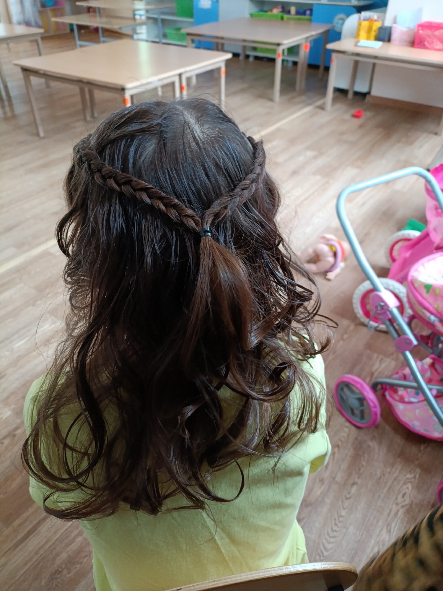 A high-angle shot shows the back of a young girl with dark, wavy hair. Her hair is braided in two sections near the crown of her head, joining together at the back and tied with a black elastic band. She is wearing a light green shirt.
The background includes a room with light brown wooden flooring. Several light-colored wooden tables and chairs are visible. A pink toy stroller and a doll are on the floor. The location is a room that appears to be a classroom.