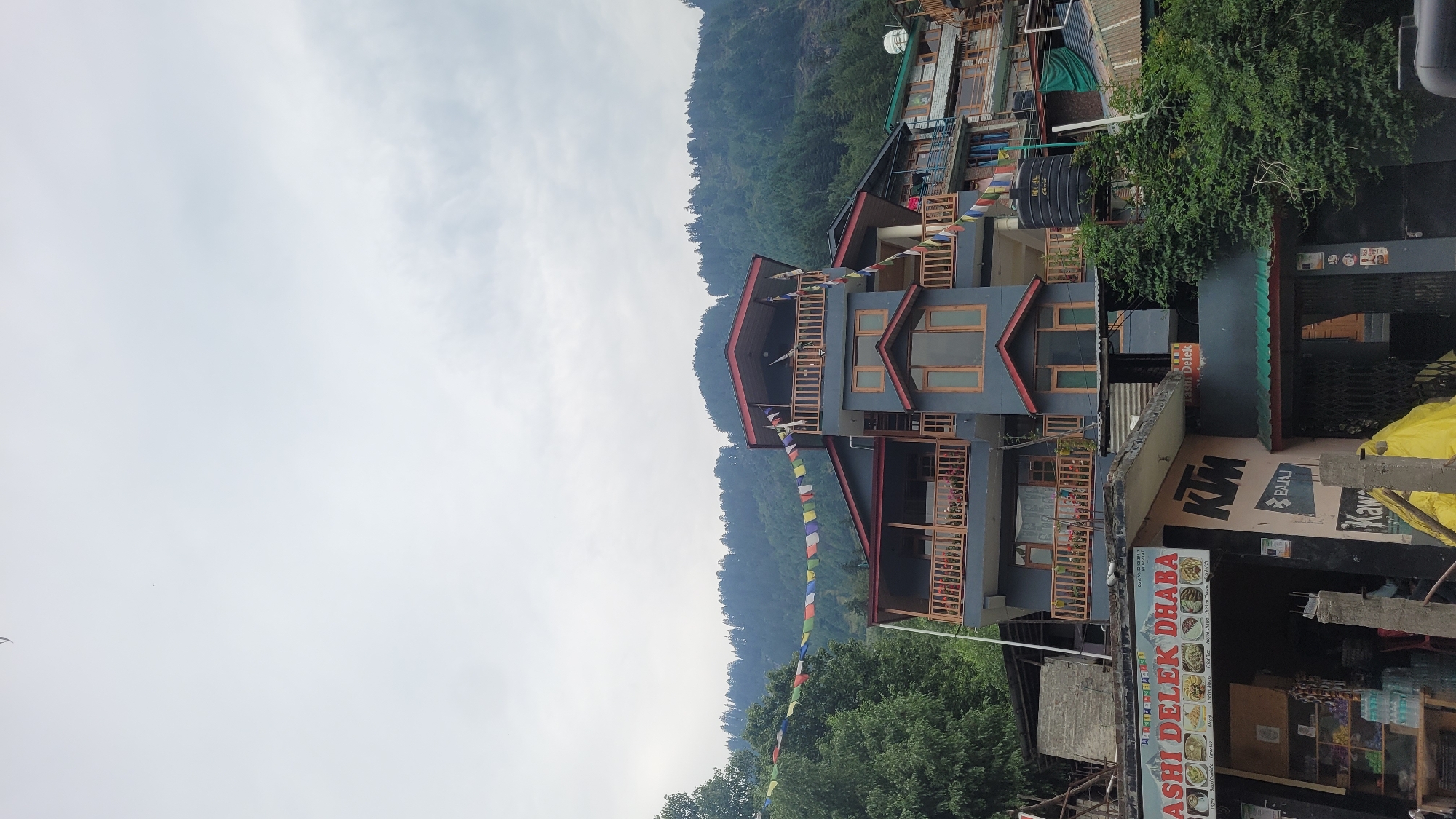 The image shows a multi-story building in Vashisht, India. The building is painted dark gray with red trim and features multiple balconies with wooden railings. Prayer flags are strung across the front of the building, visible against a cloudy sky. In the foreground, there is a shop with a sign that reads "KASHI DELEK DHABA." The shop appears to sell drinks and snacks. To the right of the shop, there is a large black water tank mounted on the side of the building. Behind the building, a densely forested mountain slope rises up. The overall impression is of a commercial building in a mountainous, possibly tourist-oriented area.