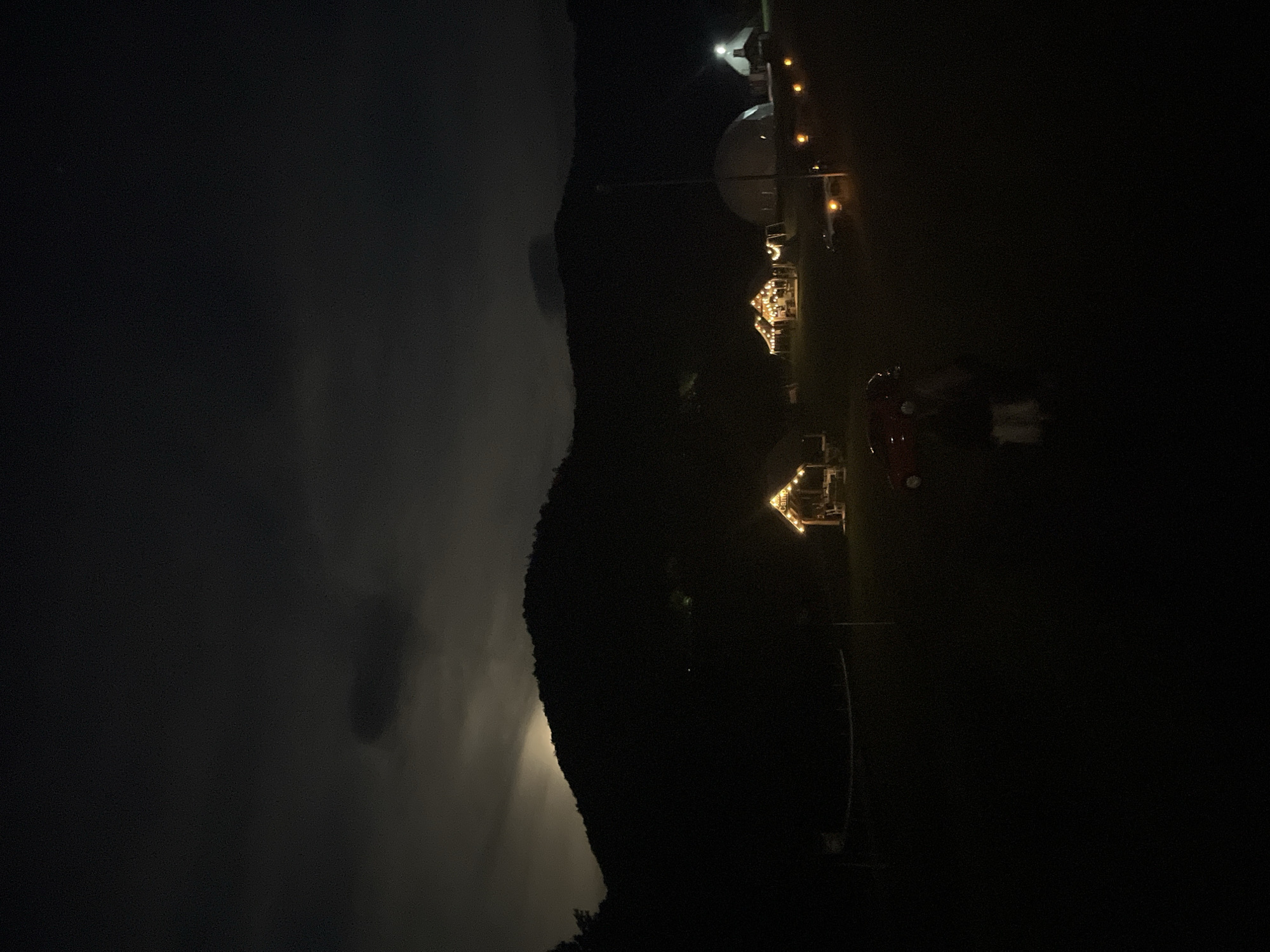 The image shows a dark, nighttime scene in Polja, Montenegro. A mountain partially obscures the left side of the frame, with visible clouds and the suggestion of moonlight breaking through. On the right side of the image are buildings or structures illuminated with yellow lights. From top to bottom, these objects can be seen: a white building with a satellite dish and a row of lights, a building with lights and foliage, a red car and two white tents, and a cabin with lights. These lights, along with faint details of the landscape, break the darkness of the image.
