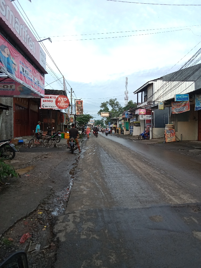 A street scene showcases a narrow road with uneven pavement in a commercial area. Multiple scooters and bicycles are parked on the side of the road. People are seen riding motorcycles in the distance. Shops line both sides of the street, adorned with signs and banners. Overhead power lines crisscross the scene. The sky is overcast, suggesting it might have recently rained or is about to rain.