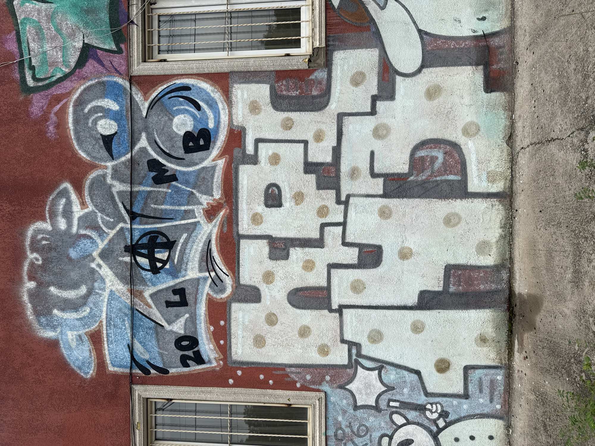 This is an exterior shot of a building with graffiti covering a significant portion of its reddish-brown wall. Two barred windows are visible, one on the left and one on the right, with similar white frames. The graffiti is colorful and complex, featuring bubble letters, abstract shapes, and what appears to be an anarchy symbol. The main text in large, white bubble letters with gray and red outlines reads "HPET". Above this, smaller graffiti elements include the numbers "20", the letter "L", a black anarchy symbol, and the letter "M", with more abstract blue and gray shapes surrounding them. The wall is textured. The ground in front of the wall is a rough, cracked concrete surface with some scattered debris and a small patch of green weeds. The overall impression is of an urban environment with street art. This specific graffiti is located on the wall of a former Soviet-era bathhouse on Meskheti Street, Tbilisi, Georgia.