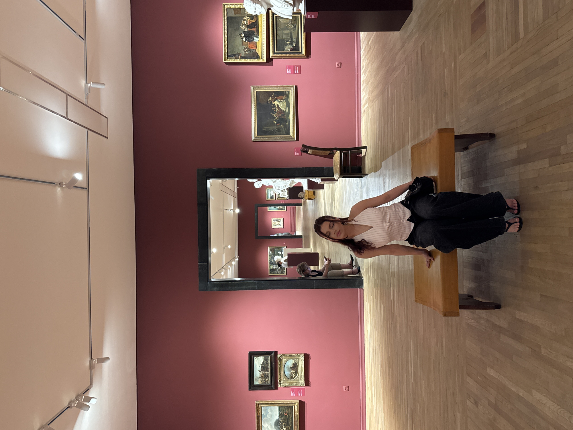 The image depicts a young woman sitting on a wooden bench in what appears to be an art museum. She is facing forward with her eyes closed and has a relaxed posture. She is wearing a white and pink striped halter top and dark pants. A black bag is slung over her shoulder, resting on the bench next to her. The museum walls are painted a deep rose color, and several framed paintings are hung on them. Through an open doorway in the background, more of the museum is visible, including other artworks and a few other patrons. The floor is made of polished light-colored wood. The lighting suggests an indoor exhibition space, with track lighting visible on the ceiling. There is no indication that this location is in Yerevan, Armenia; the scene is generic to many art museums worldwide.