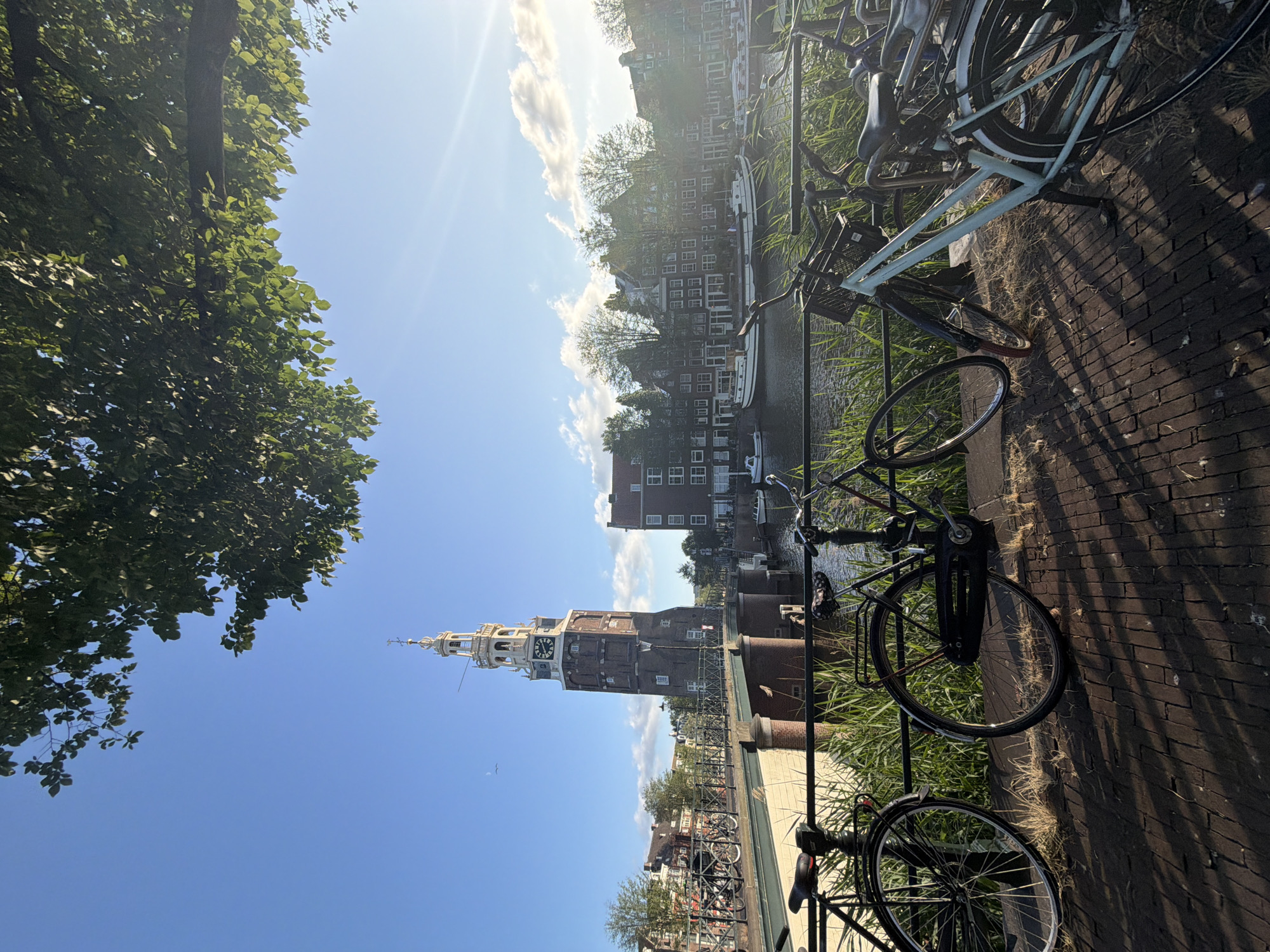 A vertical, sunlit, eye-level shot captures a canal scene in Amsterdam, Netherlands. A prominent, brick clock tower stands tall on the left side of the frame, connected to a bridge spanning the canal. Several bicycles are parked along the brick walkway in the foreground, casting long shadows. The canal itself is lined with traditional Dutch buildings and features a few houseboats. Lush green foliage frames the top of the image, with a tree branch extending into the frame from the upper right. The sky is a clear blue with scattered white clouds and a bright sun.