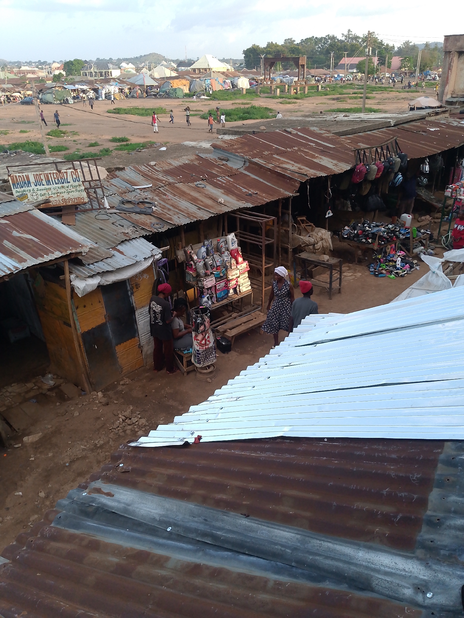 The image shows an open-air market in Bukuru, Nigeria. The market consists of multiple stalls with corrugated metal roofs, some rusty and some new. In the foreground, there are two roofs, one rusty and one newer silver. People are seen browsing and interacting at the stalls, which display various goods, including shoes, bags, and automotive parts. In one stall, a person appears to be getting their hair styled. In the background, there is a large open area with more dispersed structures and people moving about. Signage for "MADAM JOE INTERBIZ CO" is visible on one of the stalls, advertising the sale of Peugeot, Golf, and Japanese spare parts. The sky is overcast.