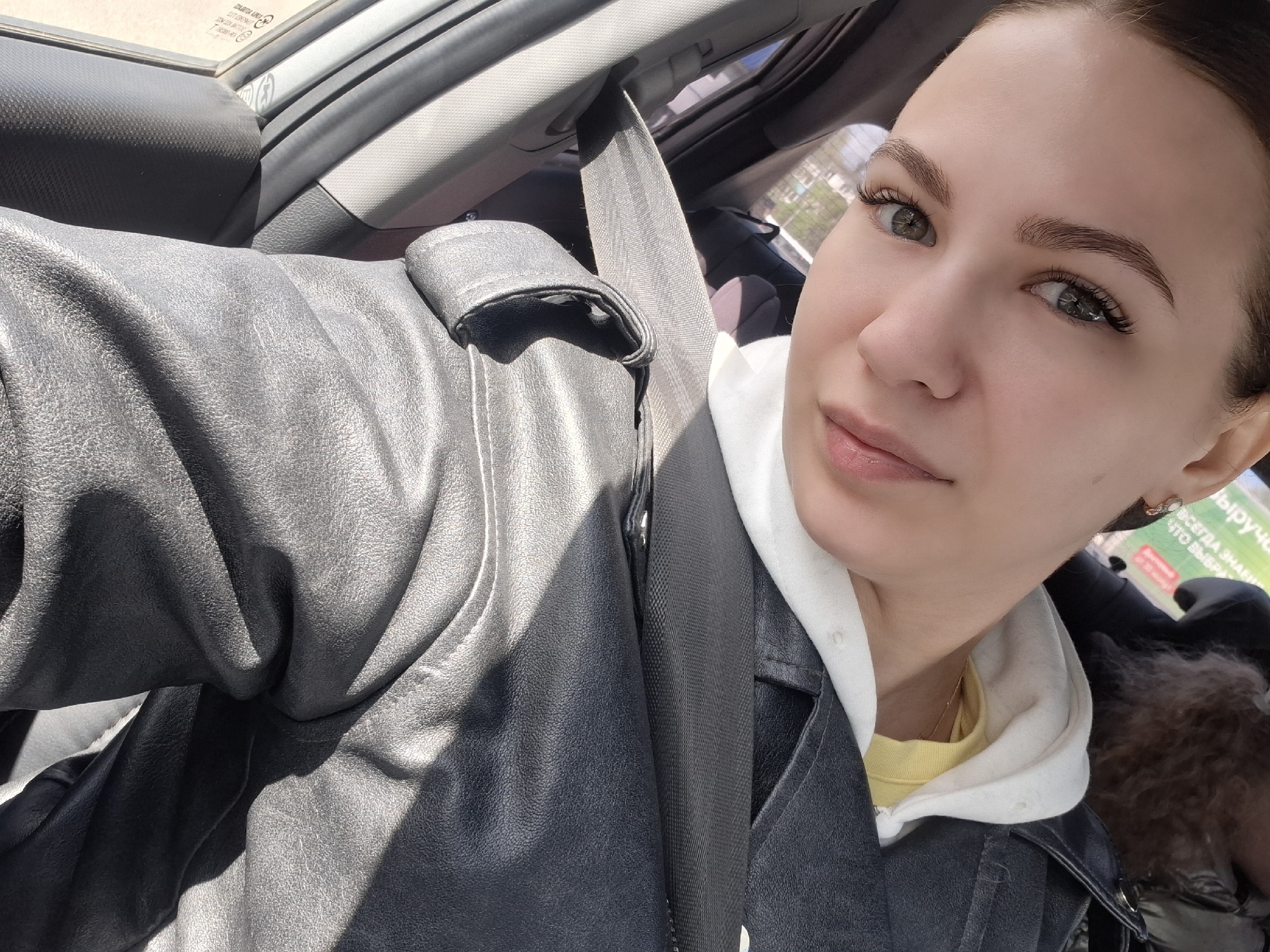 A young woman is positioned inside a car, wearing a yellow t-shirt underneath a white hoodie and a black leather jacket. A seatbelt is fastened across her torso. The car's interior is visible, including the dashboard and seats. Outside the car window, a building with text in Russian is noticeable, which provides a clue to the location. Given the clue on the building, the location is presumed to be the city of Chaikovsky, Russia.