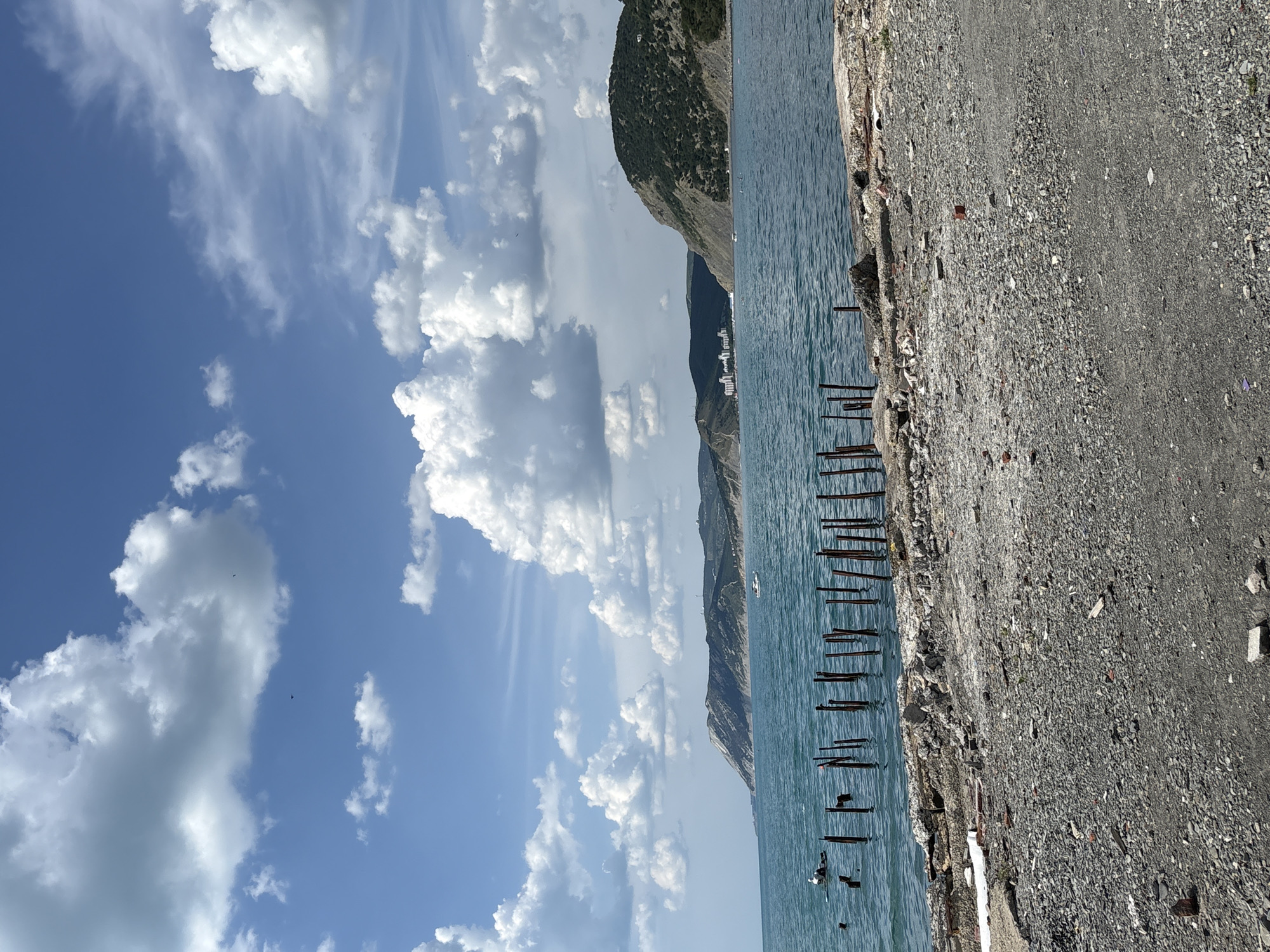 The image depicts a coastal scene in Bol'shoi Utrish, Russia, under a partly cloudy sky. The horizon features a vast expanse of turquoise-blue sea, bordered by a series of undulating green and rocky mountains and hills.
The sky is a clear blue, populated by numerous white cumulus clouds of varying sizes, with some wispy cirrus clouds also present. A few small, dark specks, likely birds, are visible in the upper sky.
In the midground, the calm sea stretches across the frame. On the left side of the water, a person wearing a white top is seated on a dark paddleboard or similar small watercraft. A white bird, consistent with a seagull, is perched on the front of the paddleboard. Further to the right, a white motorboat is visible on the water. In the far distance, on the right side of the bay, several white, multi-story buildings are situated at the base of the hills, indicating a settlement.
Protruding from the water in the immediate midground are multiple vertical, dark, rusted pilings, likely remnants of an old pier or structure. These pilings extend from the left toward the center of the frame.
The foreground consists of a rough, uneven shoreline. The ground is composed of a dense layer of grey gravel, small stones, and scattered debris including fragments of concrete, reddish brick pieces, and lighter-colored refuse. The immediate edge where the land meets the sea is formed by broken and eroded concrete structures.