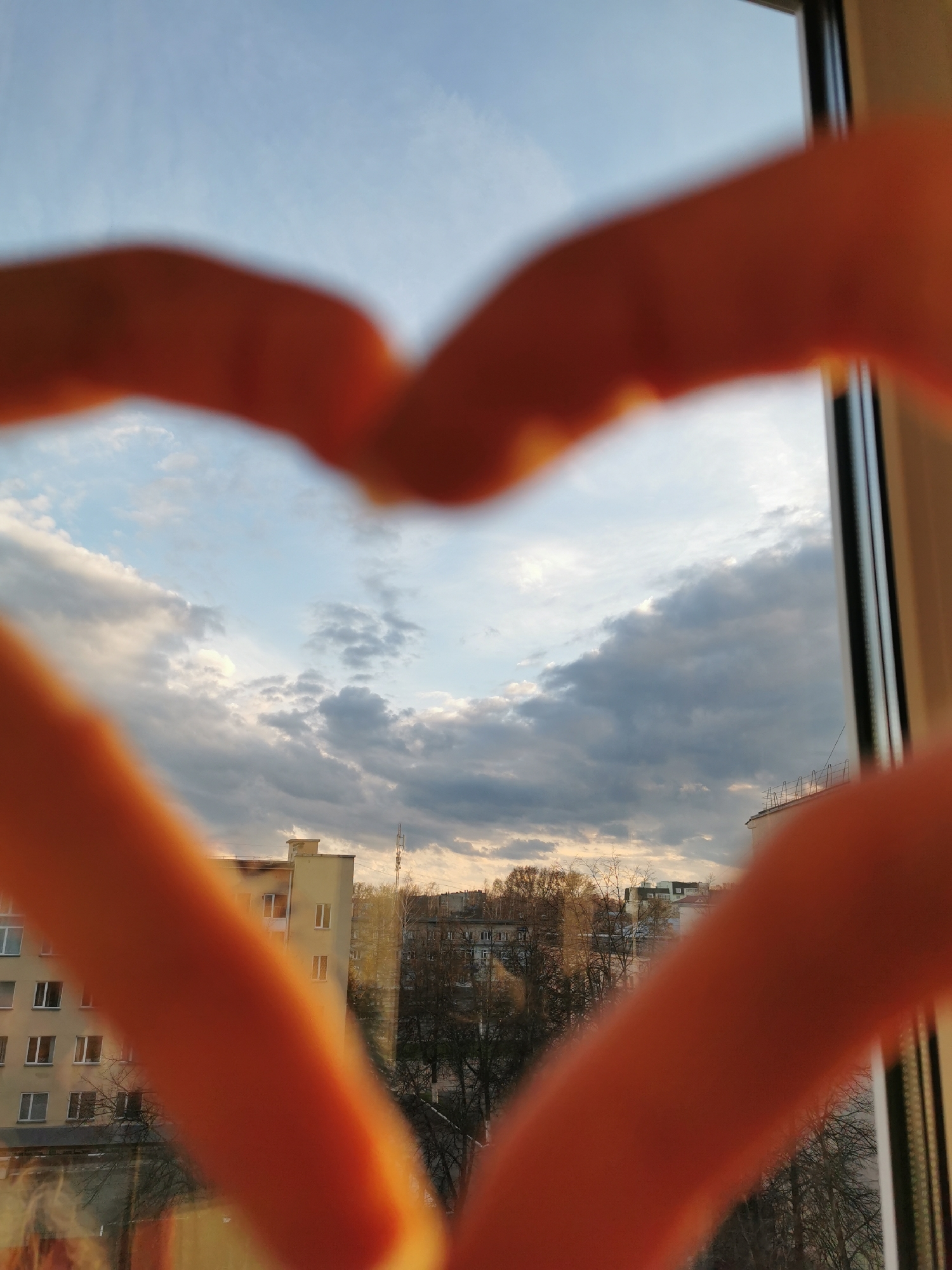 The image captures an outdoor urban or suburban landscape framed by blurred human hands forming a heart shape, seen through a window. The hands, out of focus and orange-toned due to light, occupy the foreground, with the fingertips meeting at the top center to complete the heart outline.
Beyond the hands, the view reveals a sky dominated by a mix of light blue and grey clouds, suggesting a partially overcast day. Sunlight appears to break through in places, illuminating some clouds from behind. In the middle ground, several multi-story buildings are visible. One prominent light-colored building, possibly residential, stands on the left, with numerous windows. Another building, slightly darker and partially obscured by trees, can be seen further back. Bare tree branches, characteristic of late autumn, winter, or early spring, fill the space between the buildings and the distant background. A slender antenna or communication tower rises above the buildings on the right side. A window frame is visible along the right edge of the image.
The light quality, with a mix of sun and clouds, suggests it might be late afternoon or early evening. The overall impression conveyed by the heart-shaped frame is one of affection or appreciation for the view. There is no visible text in the image.