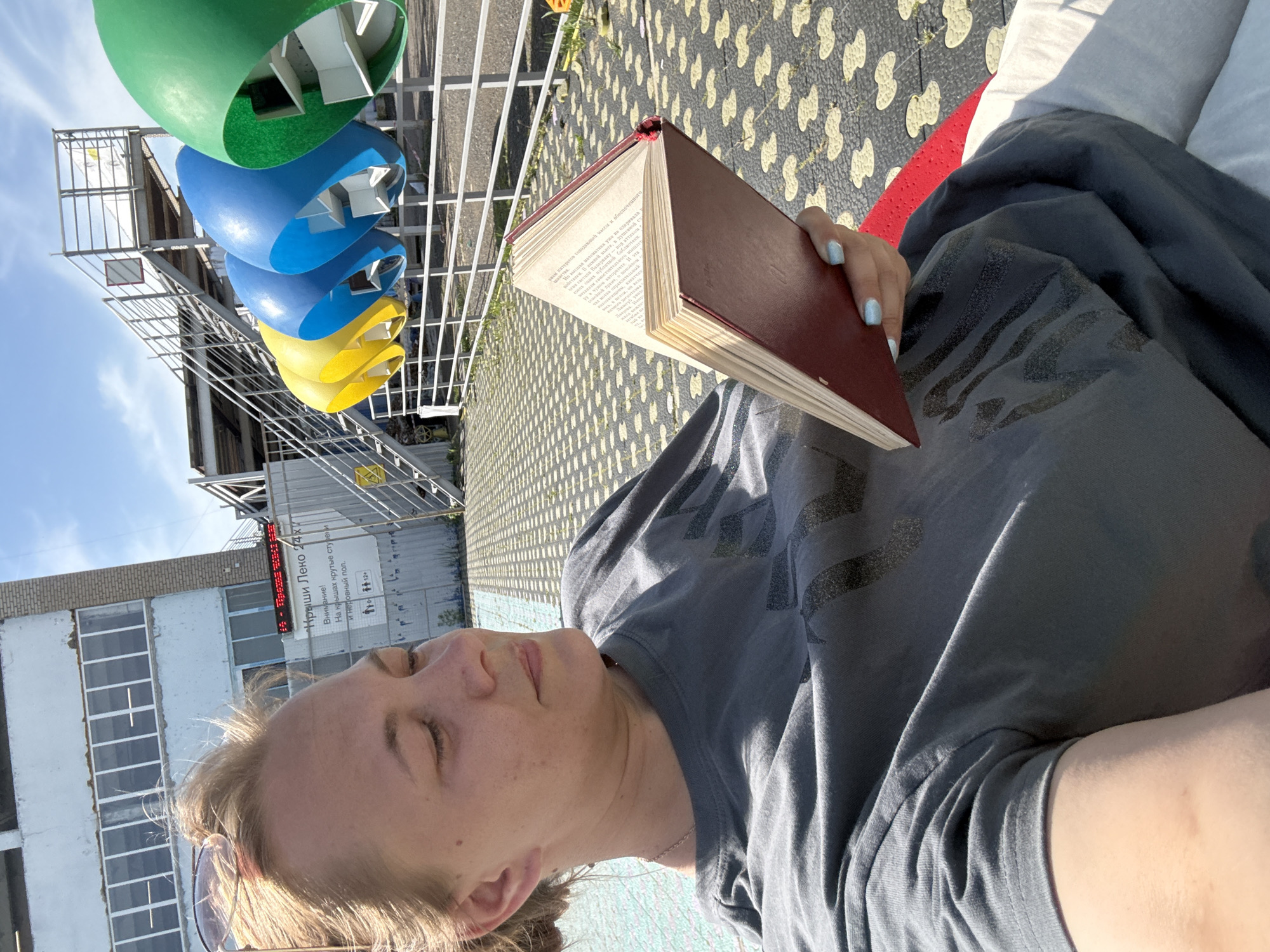 A woman is lying down, reading a book, outdoors in what appears to be the city of Sin’kovo, Russia. The book has a red cover and open pages. The woman has blonde hair and is wearing a gray T-shirt. The location is a public space; to the left is a multi-colored sculpture of what appear to be green, blue, and yellow funnels. There is a building with a sign containing Russian text that reads "Kryshi Leko 24x7". The ground is patterned.