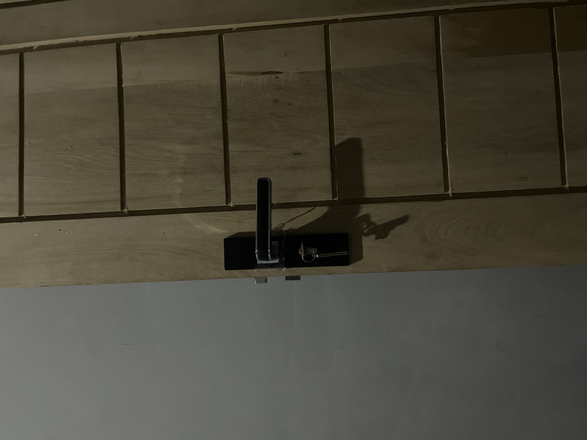 The image shows a close-up view of a wooden door with a black door handle and keys. The door is divided into horizontal panels. To the left of the door is a plain gray wall. The scene appears to be indoors, possibly in a residential or commercial building. The lighting suggests it is nighttime or a dimly lit interior. There is no indication of the specific location in Kebumen, Indonesia, beyond what can be inferred from the general appearance of a door and wall.