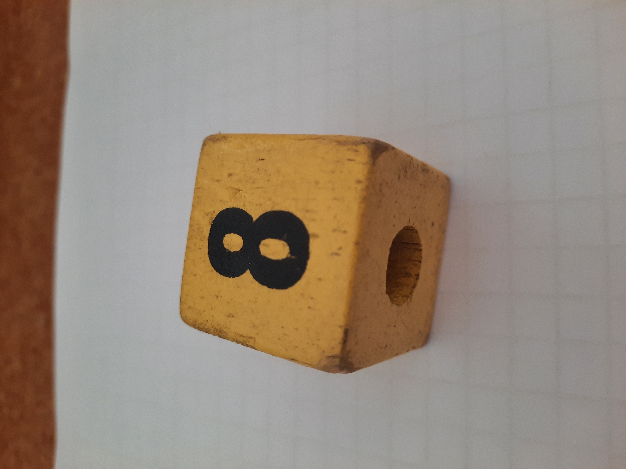 A yellow wooden cube with a black number "8" on one face and a hole on an adjacent face rests on a white grid-lined surface. The surface is set against a blurred background of a brown wooden table. The image appears to be taken in a domestic setting and there is no indication of it being in Lhokseumawe, Indonesia.