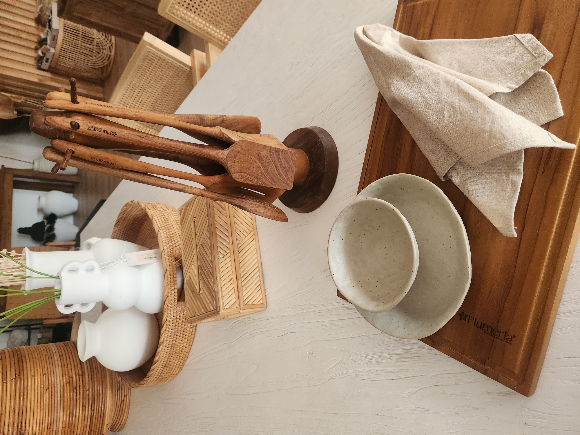 The image displays a well-lit, close-up, eye-level shot of a white wooden table, presenting a collection of home decor and kitchenware. The scene is arranged in a visually appealing, staged manner, suggesting a retail or domestic setting. In the upper left, a large, woven rattan basket serves as a base for two white ceramic vases. The vase on the left is bulbous with a narrow neck, while the one on the right is more ornate, featuring two curved handles. A tag is attached to the right vase. In the foreground, a wooden cutting board holds a folded light-beige linen napkin and a stack of two speckled ceramic dishes, one a shallow bowl atop a plate. To the left of the cutting board, a rectangular rattan tissue box with a patterned lid is placed on the table. Dominating the center and right of the image is a wooden utensil holder with multiple wooden spatulas and spoons, their handles engraved with the "Plumeria" logo. The background is softly blurred, showing glimpses of furniture and decor, including more rattan elements and wooden structures. The overall aesthetic leans towards a natural, bohemian, or coastal style. The scene is static, with no visible people or actions. The lighting is soft and natural, highlighting the textures and muted colors of the objects. The location is specified as Maitland, United States of America, though the image itself provides no direct visual cues to confirm this.
