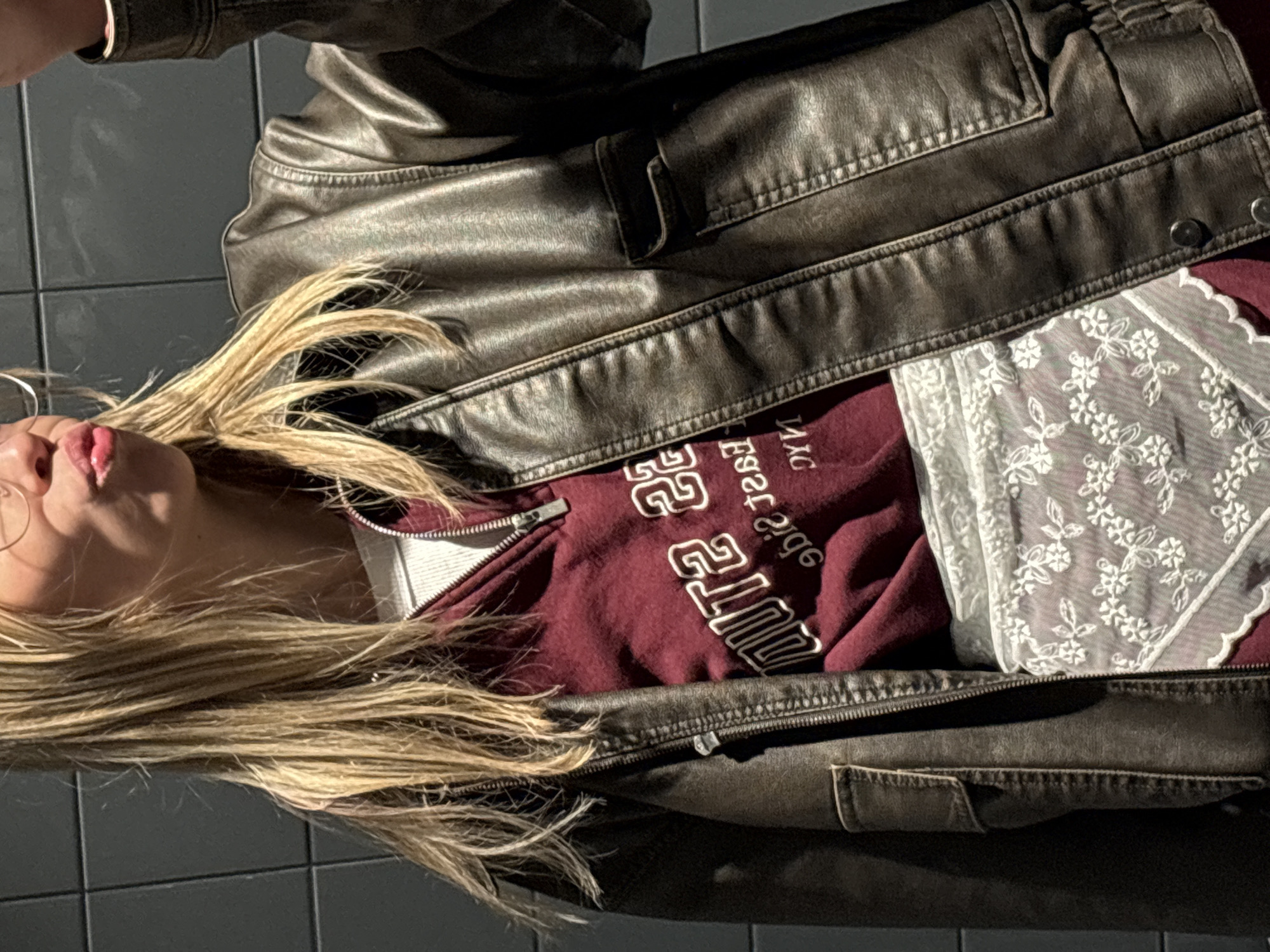 A blonde woman is lying on a grey tiled floor. She is wearing glasses and a brown leather jacket over a maroon zip-up hoodie with white lettering. She is also wearing a white lace top. The location is unknown and cannot be determined to be Krasnoyarsk, Russia from the given image.
