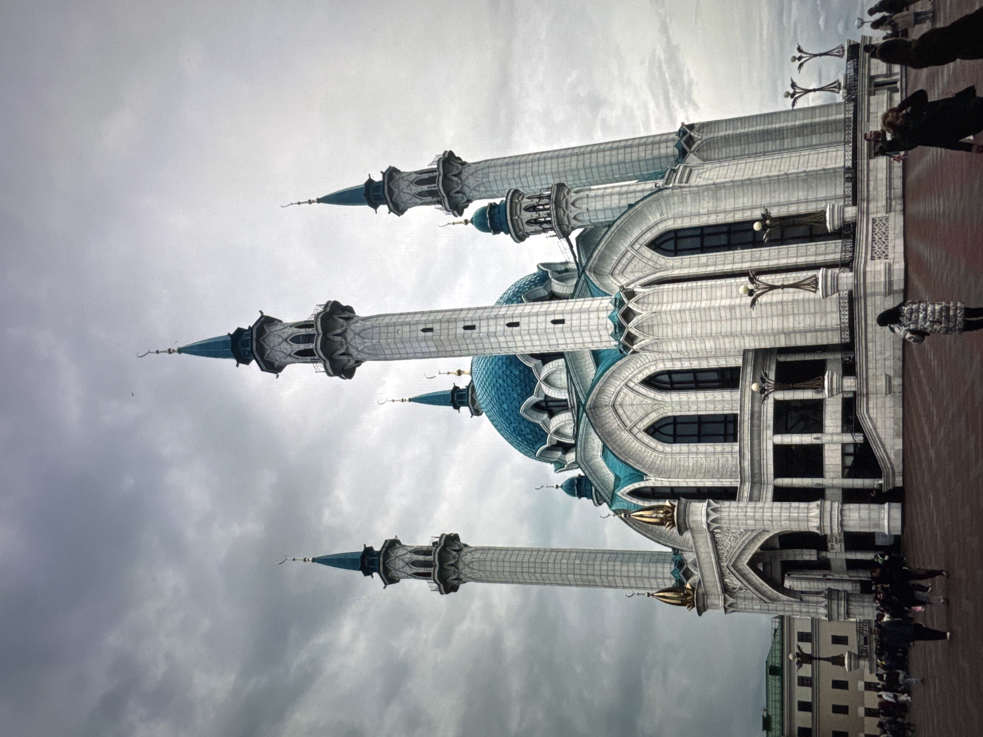 A low-angle, rotated shot reveals the Kul Sharif Mosque in Новинки, Russia, set against an overcast sky. The mosque exhibits a light-colored facade, complemented by its prominent blue dome and minarets. A group of people are visible in the lower right quadrant of the photo, standing on a red-brick paved area adjacent to the mosque. In the extreme lower right, a modern building can be seen in the background.