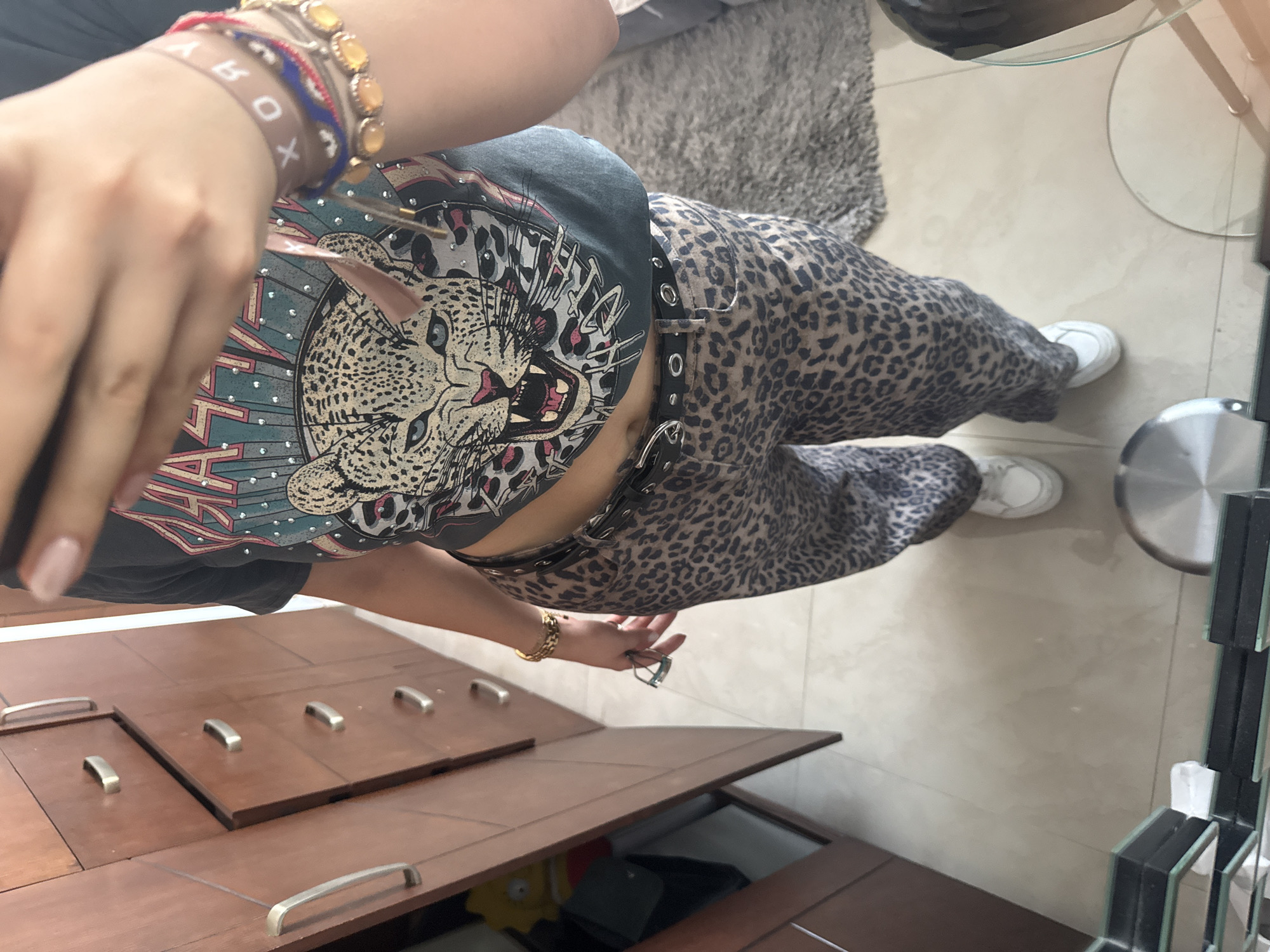 A person is standing indoors wearing a gray t-shirt with a leopard print design, leopard-print pants, and white sneakers. They have bracelets on their left wrist and are holding eyeglasses in their right hand. The setting appears to be a home, with furniture including a wooden dresser or cabinet and a glass-topped table with a gray rug nearby. A decorative mirror is visible on the right side of the frame. There is no discernible evidence to indicate the city of Santa Clara Ocoyucan, Mexico, is the location.