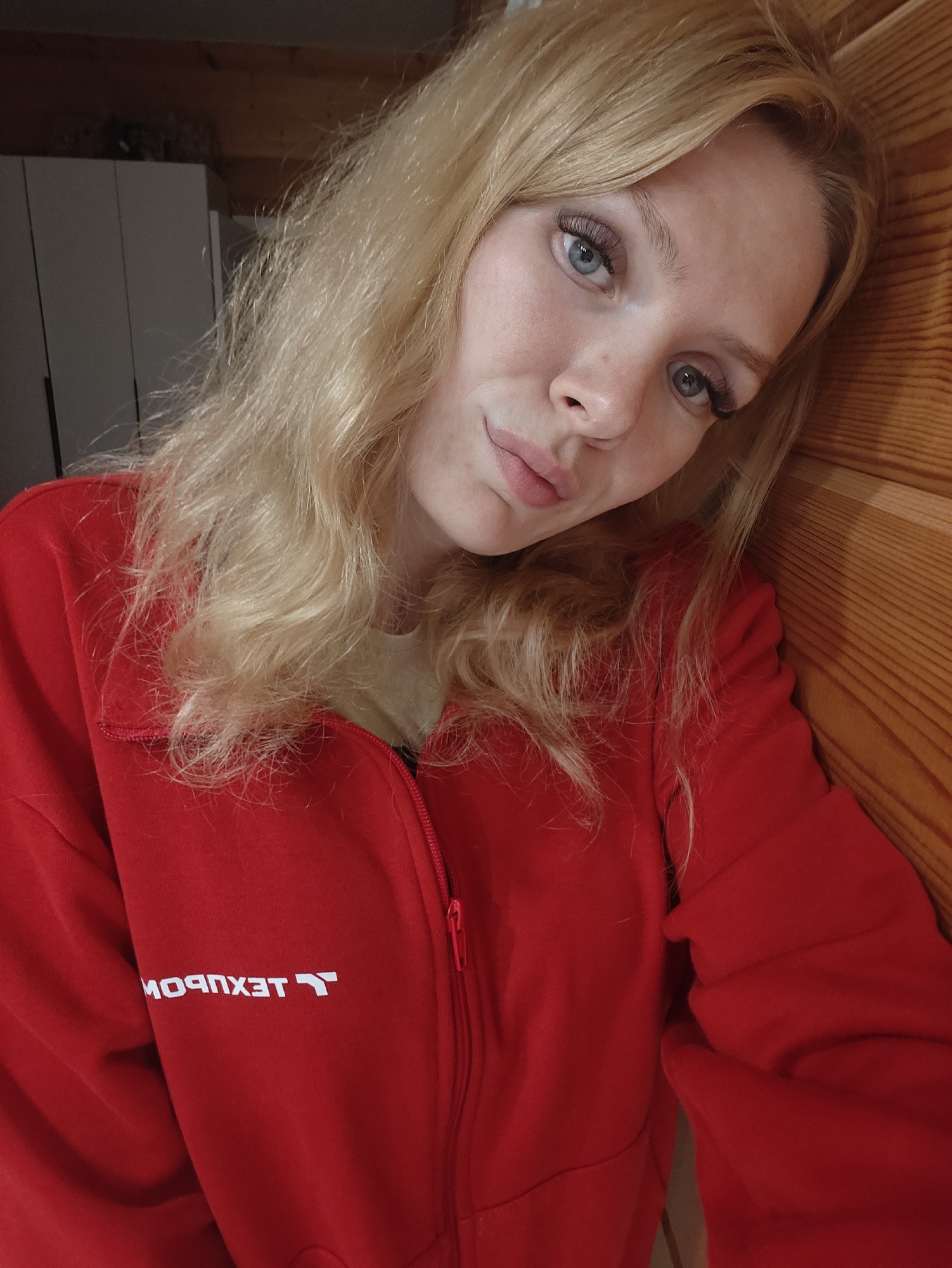 A young woman with blonde hair and blue eyes, wearing a red zip-up jacket with a white logo that reads "TEKMPOXET" in reverse, leans against a wooden surface. Her hair is styled loosely, framing her face. She is looking towards the camera with a subtle smile. The background suggests an indoor setting, possibly a room with wooden paneling and a glimpse of a white closet or cabinet. There is no indication of the location being in the city of Русский Вожой, Russia.