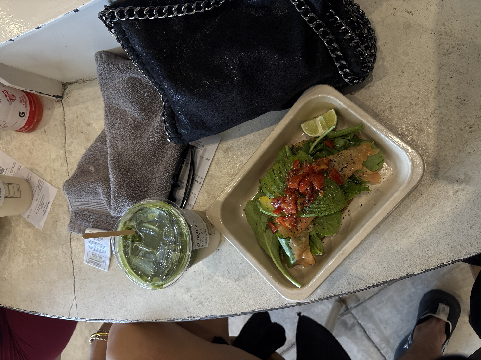 An overhead shot captures a casual dining experience, likely within a cafe in Puebla, Mexico, as indicated by visible text. The central elements rest on a light-colored, textured table, possibly concrete, featuring visible cracks.
A person is partially visible at the bottom of the frame, showing a knee in a red garment, an arm adorned with a bracelet, and a leg/foot in a dark shoe. On the table, a stylish black handbag with a prominent silver chain strap sits beside a grey towel.
The main food item is presented in a rectangular, light-colored dish, featuring an open-faced meal. This dish includes sliced avocado, layers of smoked salmon, green leafy vegetables (likely spinach or arugula), diced red tomatoes, and a fresh lime wedge for garnish, all seasoned with black pepper. A clear plastic cup contains a green iced drink, identified by its label as "21 SANTIAGO GREEN ME LATTE," with a wooden stirring stick.
Other items on the table include a white paper cup, a red and white plastic bottle with "G" and "FRUTAS TROPICALES" printed on it, and eyeglasses resting on the towel. Several paper receipts or labels are scattered across the table, one explicitly showing "PUE" (Puebla) and monetary figures like "$100.00". A small white tag with "OEKO-TEX®" is also visible. The scene suggests a relaxed moment enjoying a meal and beverage.