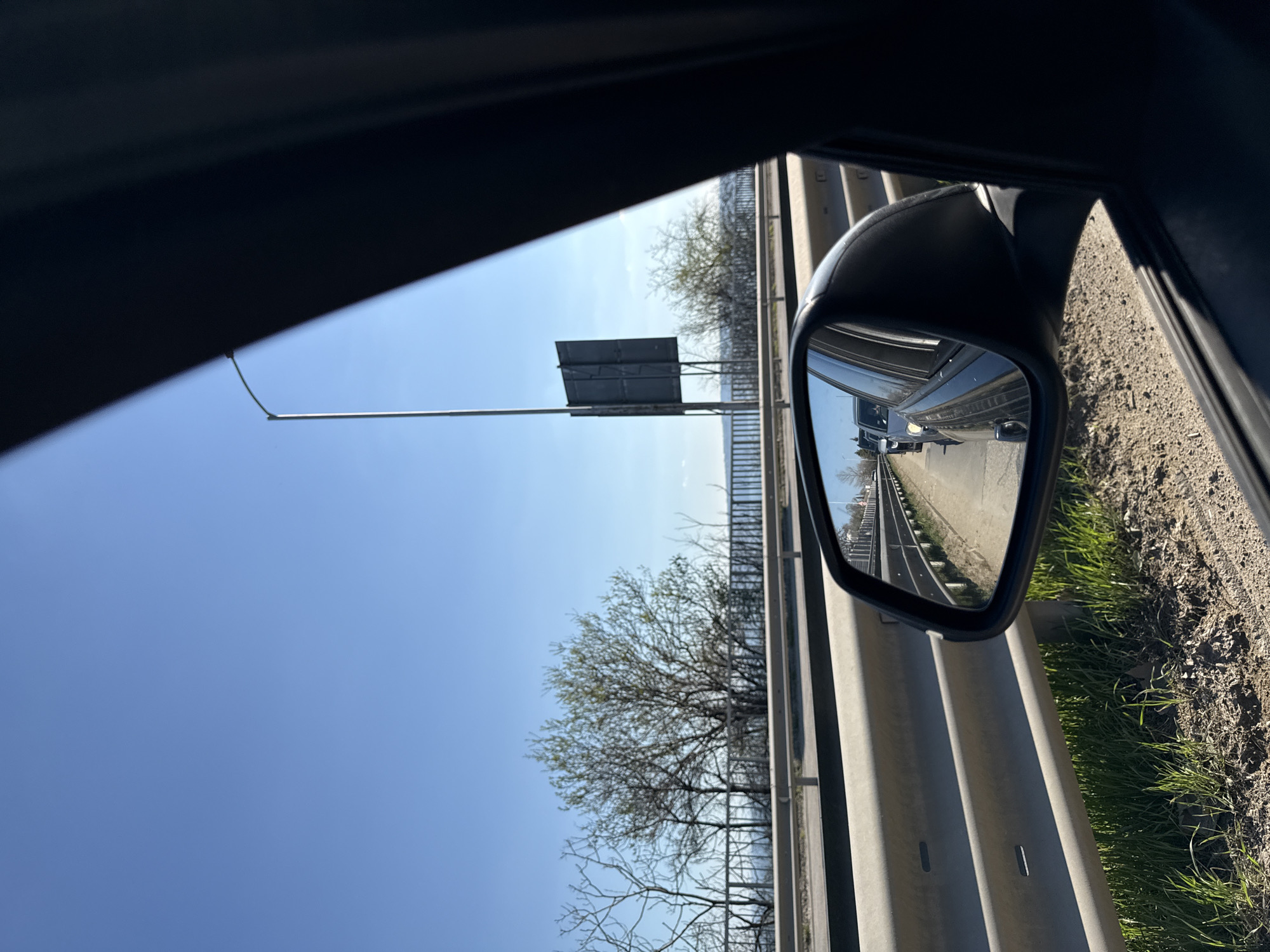 The image depicts a view from inside a moving vehicle, likely a car. The focus is on the side mirror, which reflects the adjacent lane of a highway. The mirror shows several vehicles traveling in the same direction.
Outside the car, the highway is bordered by a metal guardrail and a grassy verge. Beyond the guardrail, a row of trees lines the road. A tall signpost is visible on the left side of the image, extending above the trees. The sky is clear and blue, indicating a sunny day.
Given the details, it is likely that the image was taken on a highway near Varna, Bulgaria. Highways in the region are typically bordered by metal guardrails, and the presence of vegetation aligns with the Bulgarian landscape. The signpost may indicate a highway exit or road marker. Without additional information it is difficult to pinpoint the precise location.