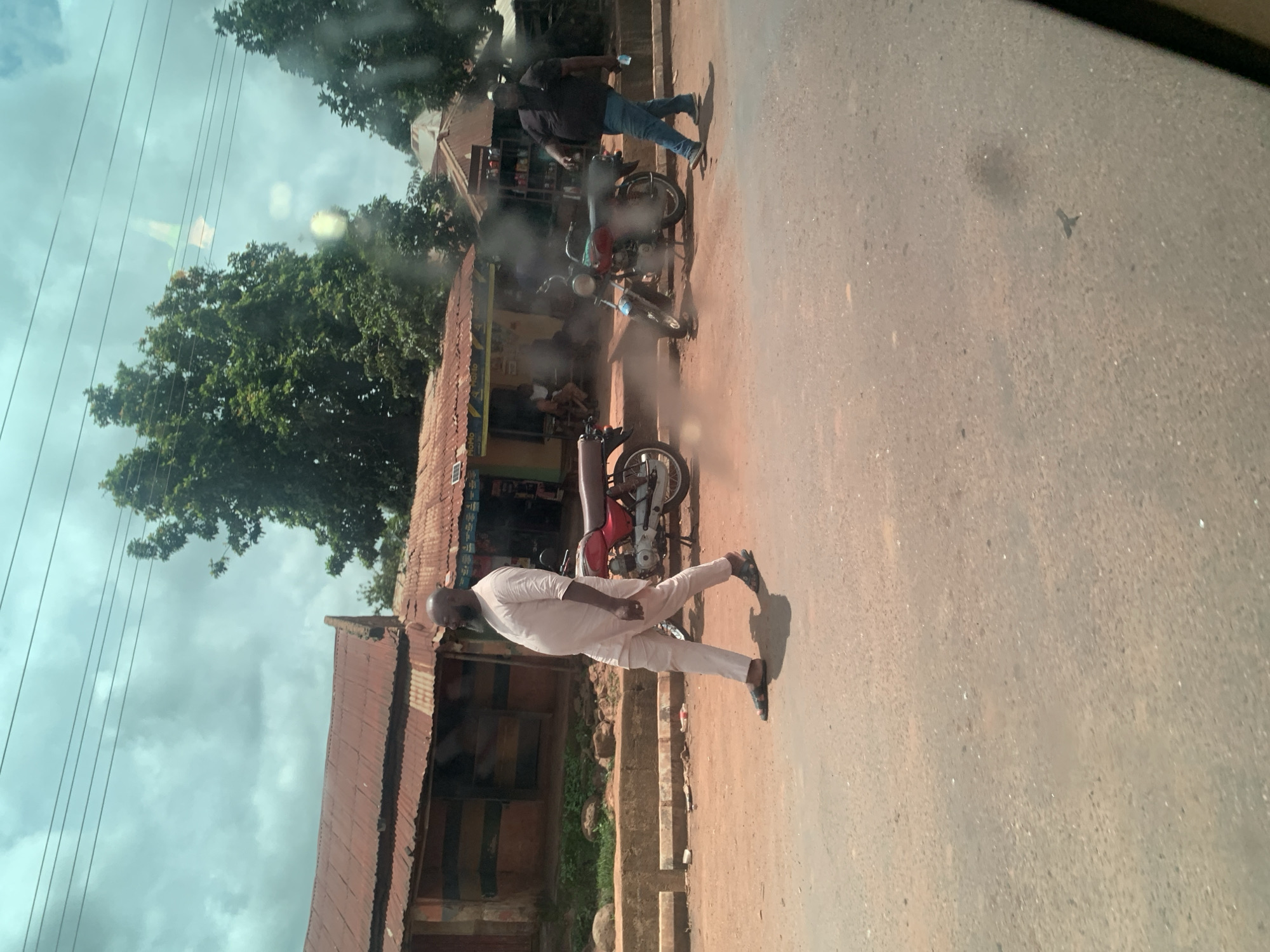 A man in a light-colored traditional outfit walks on a dirt road in Hoss, Nigeria, with a red motorcycle parked beside a corrugated iron building. Another man in a dark t-shirt and jeans walks further down the road, holding a bottle. The scene is characterized by dusty, unpaved roads, simple storefronts with metal roofs, and lush green trees in the background under a cloudy sky. Power lines are visible overhead, suggesting an urban or semi-urban environment.