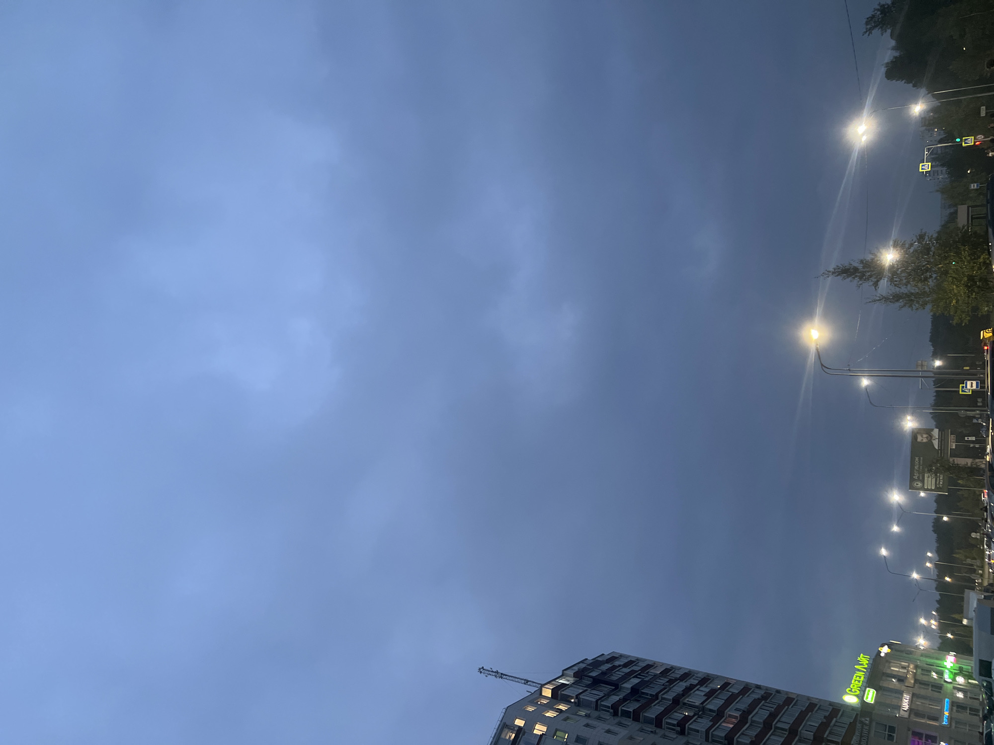 The image depicts a street scene in Surgut, Russia, under a cloudy, twilight sky. A tall, modern apartment building with illuminated windows is visible on the left, featuring a prominent antenna on its roof. Below the building, a commercial facade displays signage, including "GREEN FAIRT" in green neon letters. Streetlights illuminate a busy road with multiple lanes of traffic, although no specific vehicles are clearly distinguishable. In the background, a line of trees borders the road, and a billboard with a portrait of a man is mounted. The overall atmosphere is dim, suggesting it is either early morning or evening.